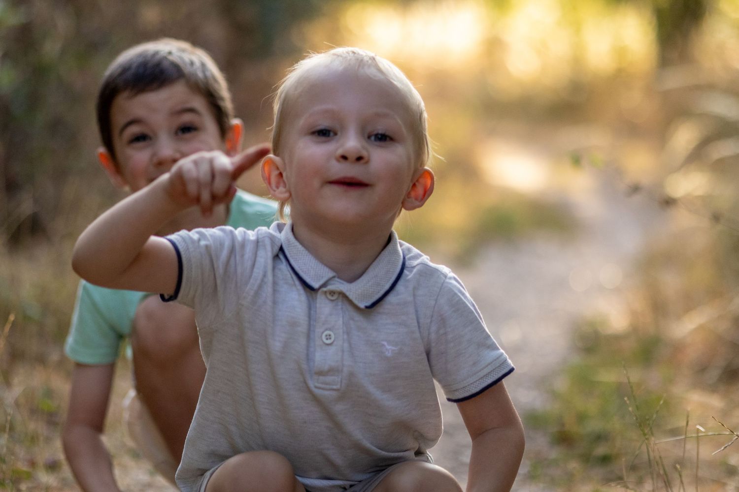 Two young boys sitting outdoors on a path, one pointing while the other looks up curiously.