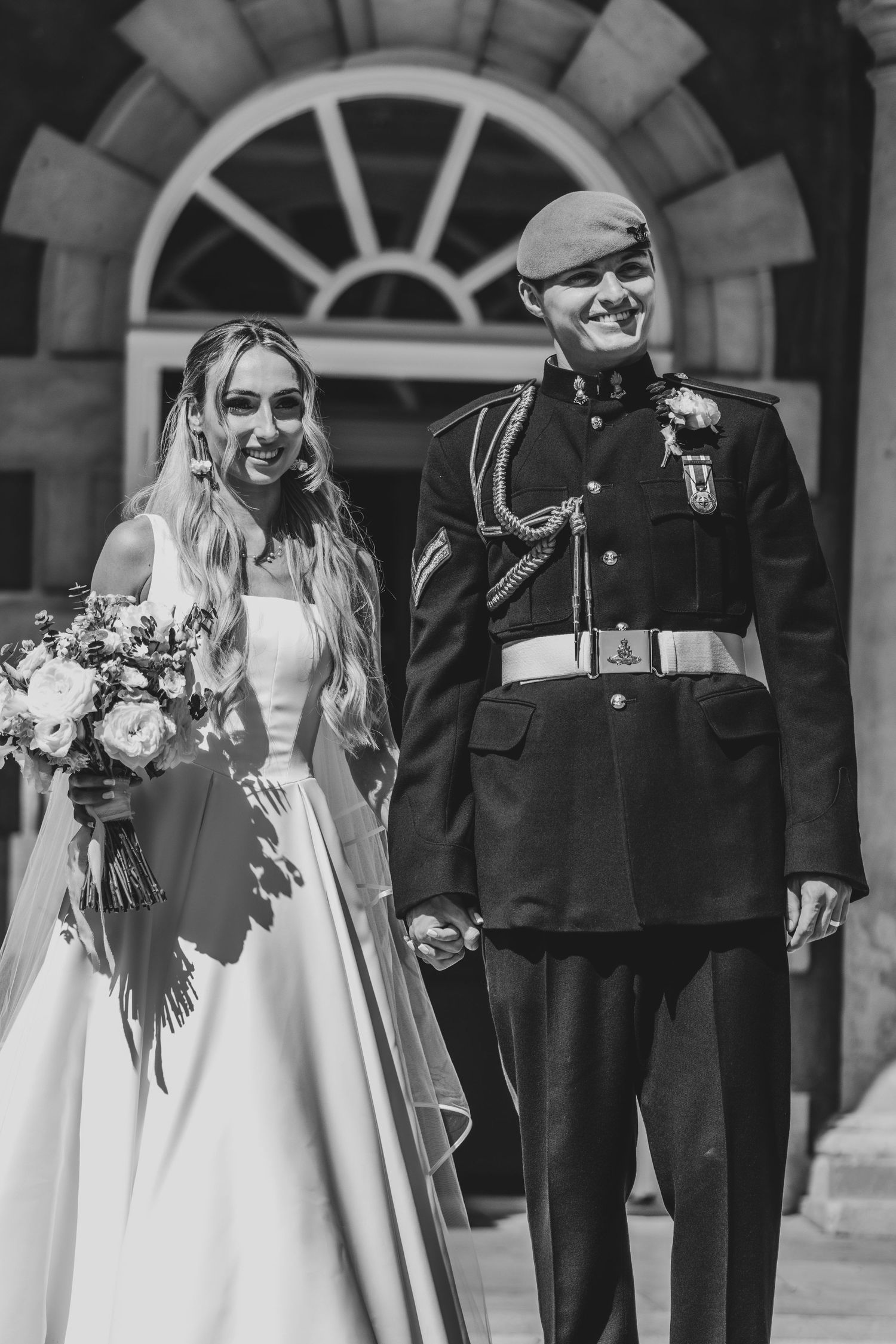 Black and white photo of smiling newlyweds, bride in white gown and groom in military uniform holding hands outside.