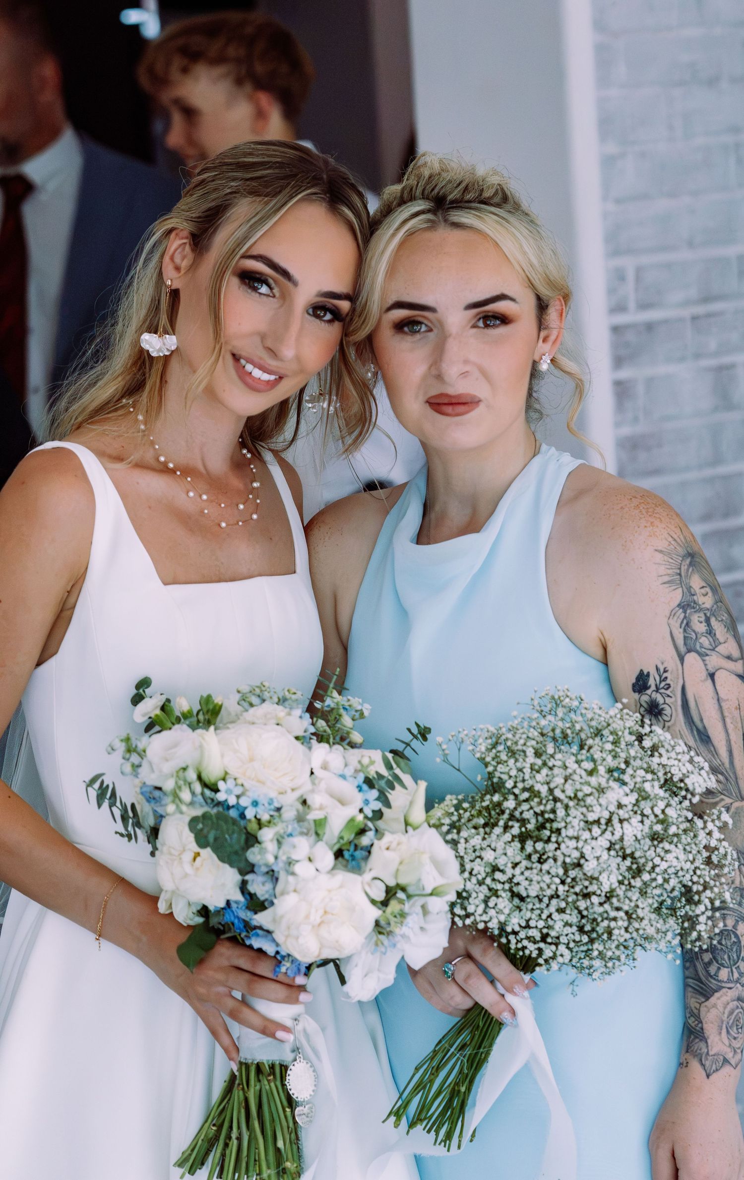 Bride holding blue and white bouquet poses with tattooed bridesmaid carrying baby's breath bouquet.