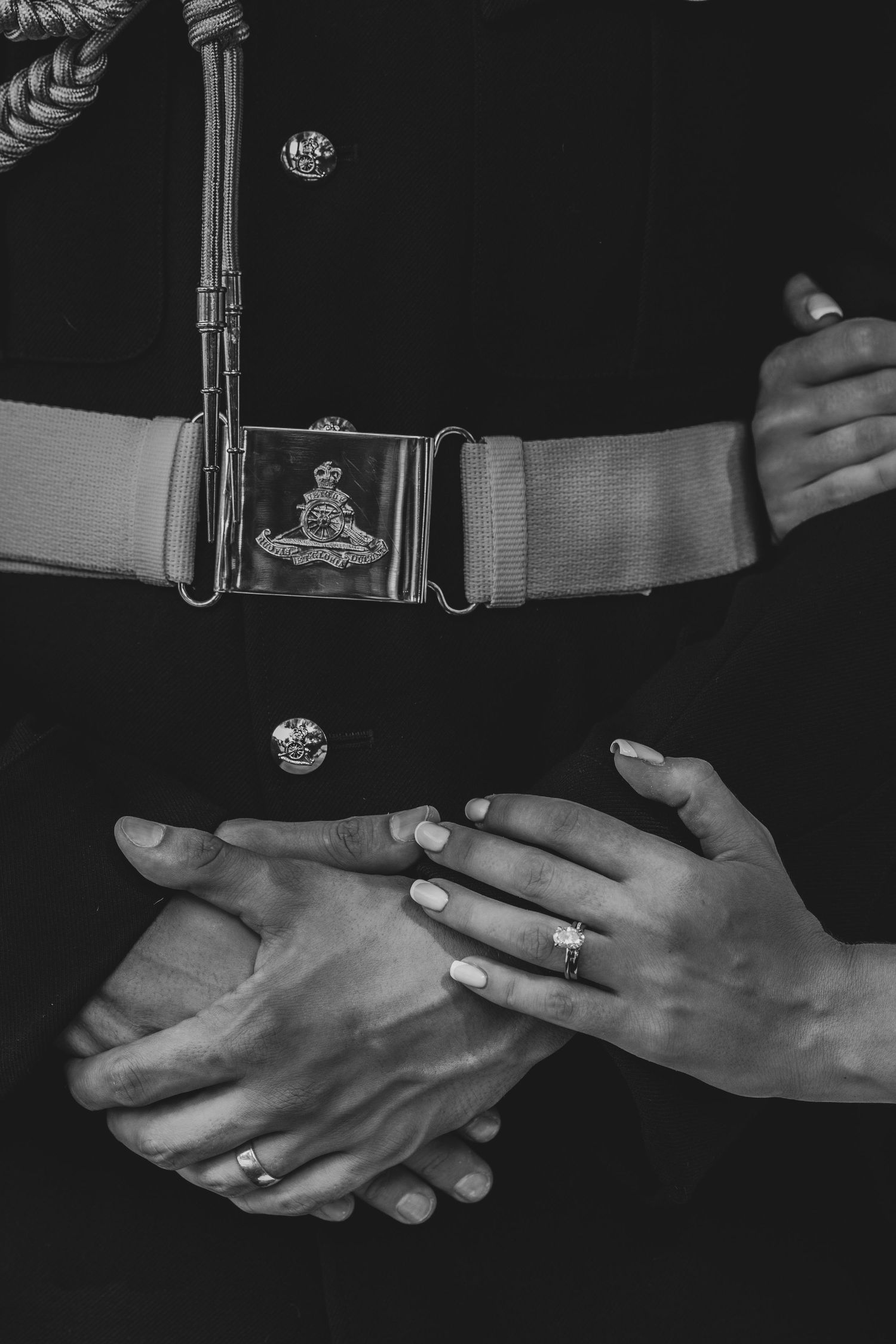Black and white close-up of clasped hands showing engagement ring beside military uniform with royal crest belt buckle.
