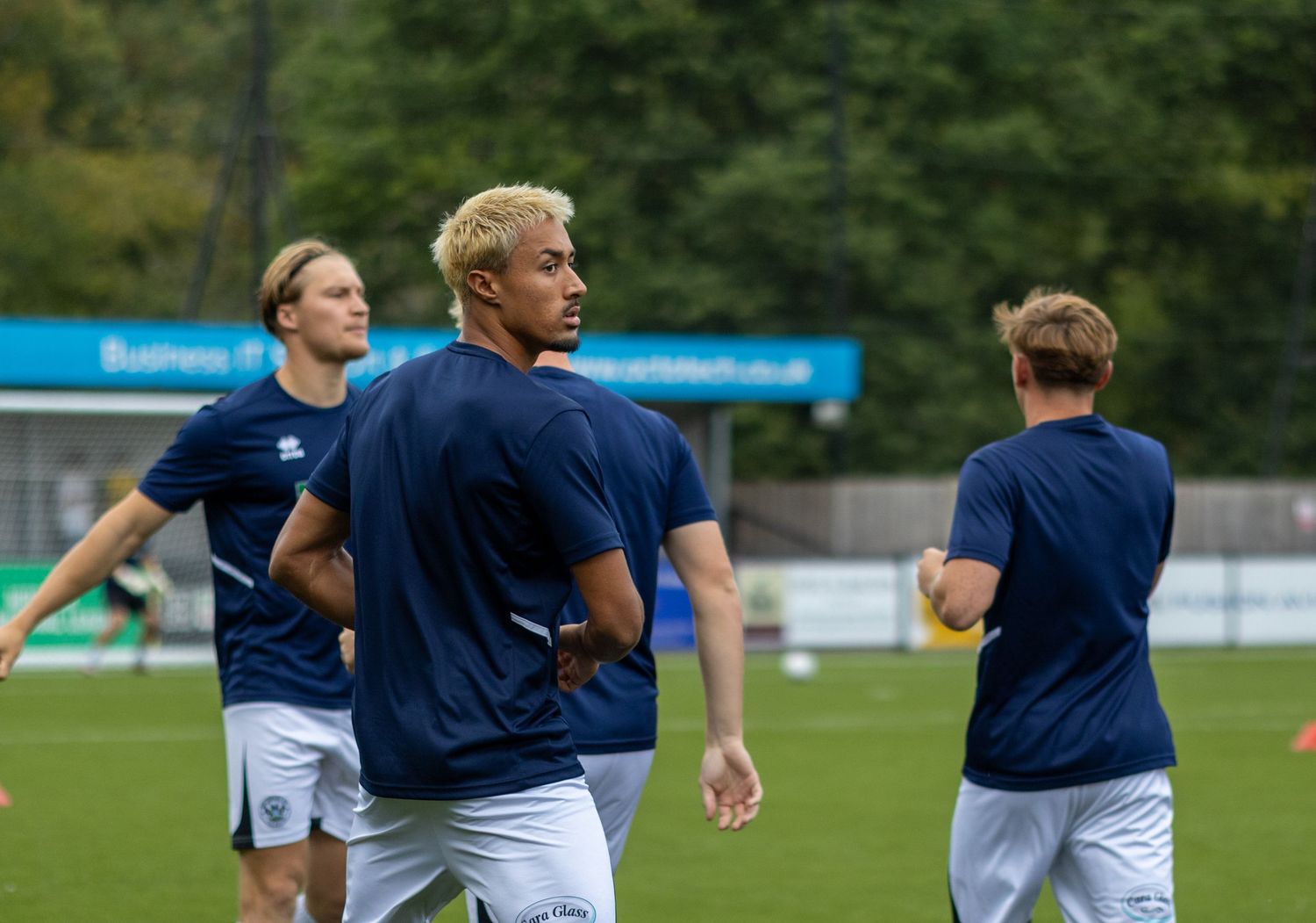 Football players in navy Errea training kit warming up on green pitch before a match.