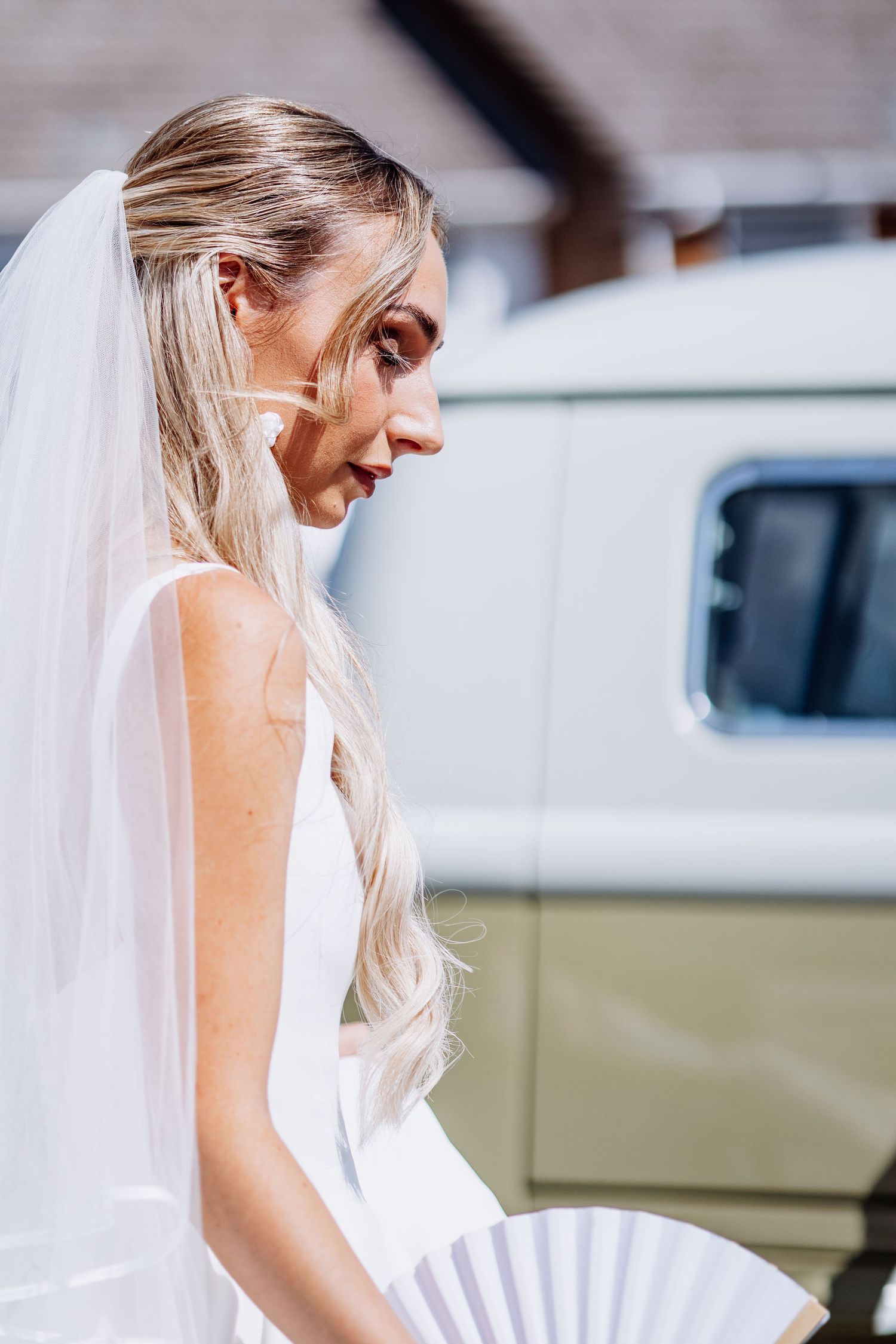 Elegant blonde bride in white dress holding a fan, wearing a veil, standing outdoors by a vintage vehicle.