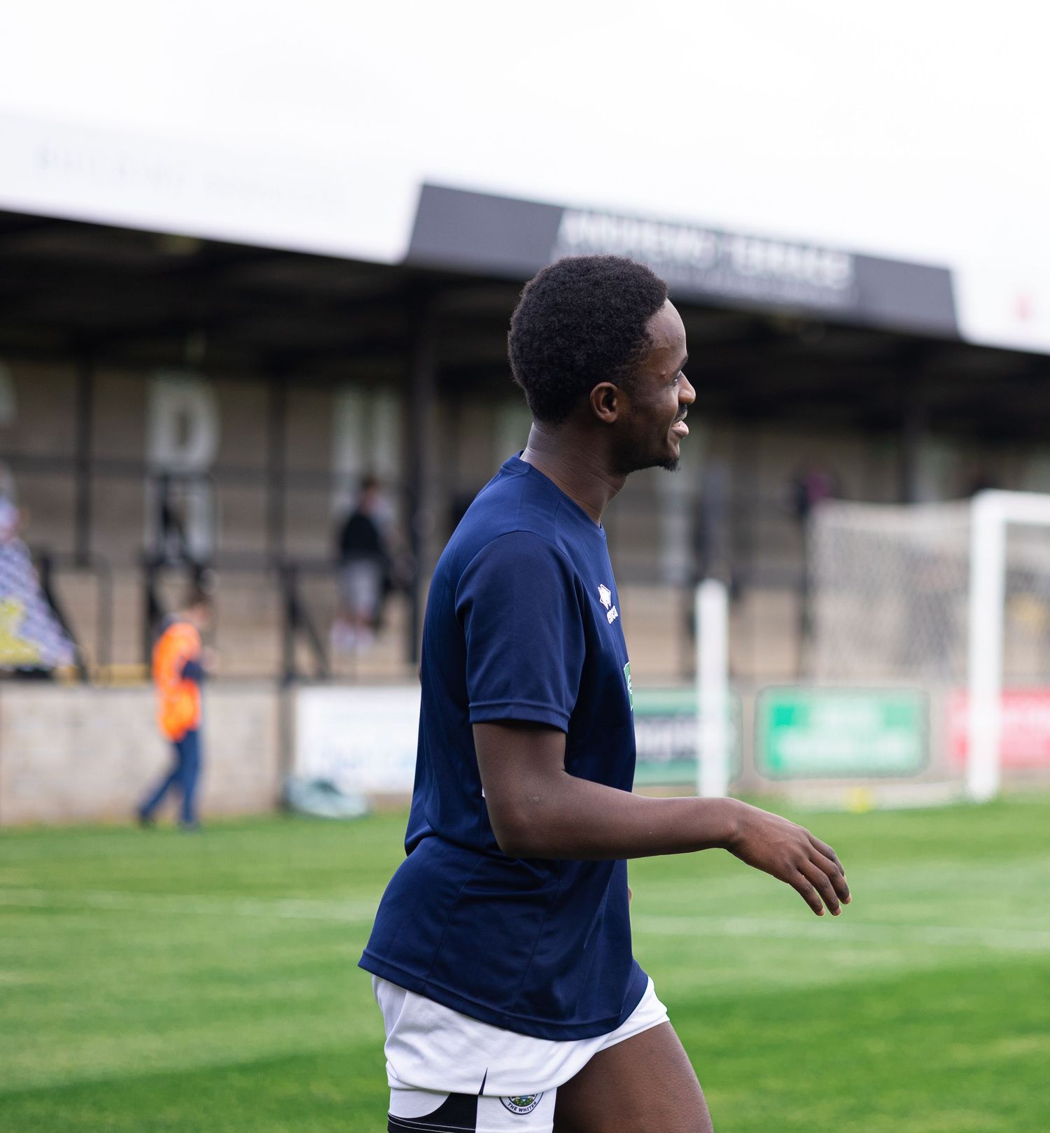 A young male soccer player in a navy blue kit smiles while jogging on a grass pitch during a match.