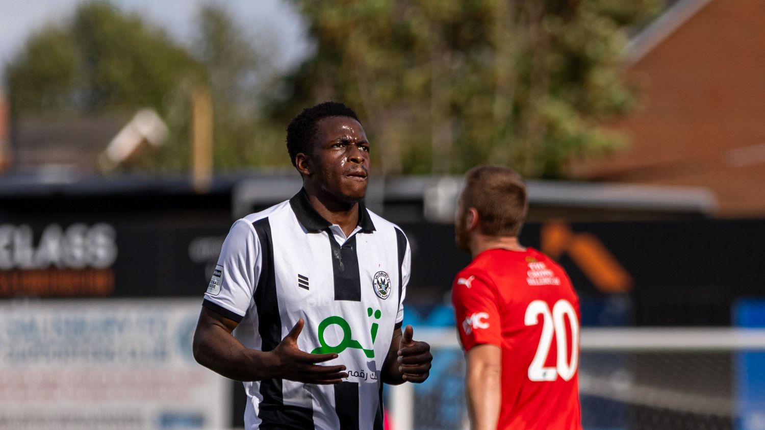 Two football players on a sunny pitch, one in black and white kit, another in red numbered 20.