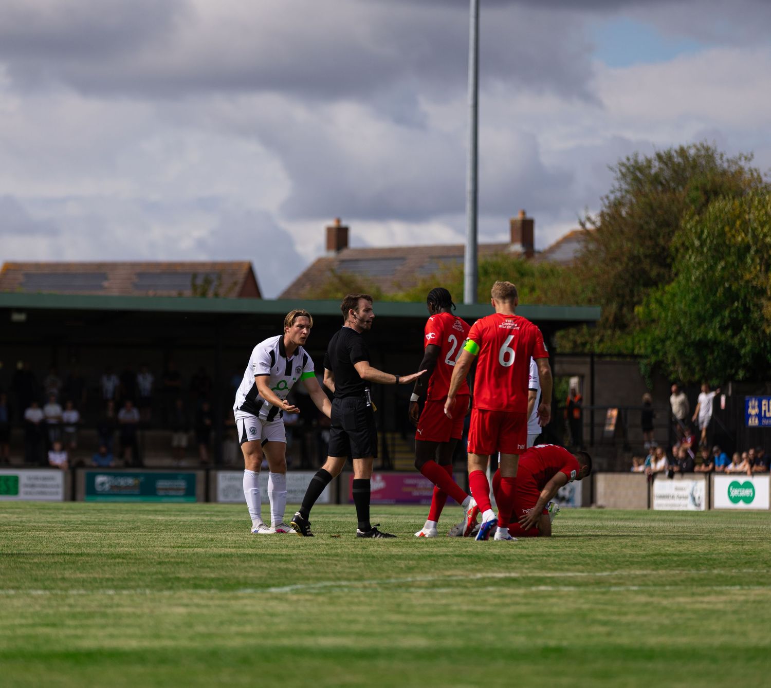 Football match scene with referee and players on green pitch, red and white kits, spectators in background.