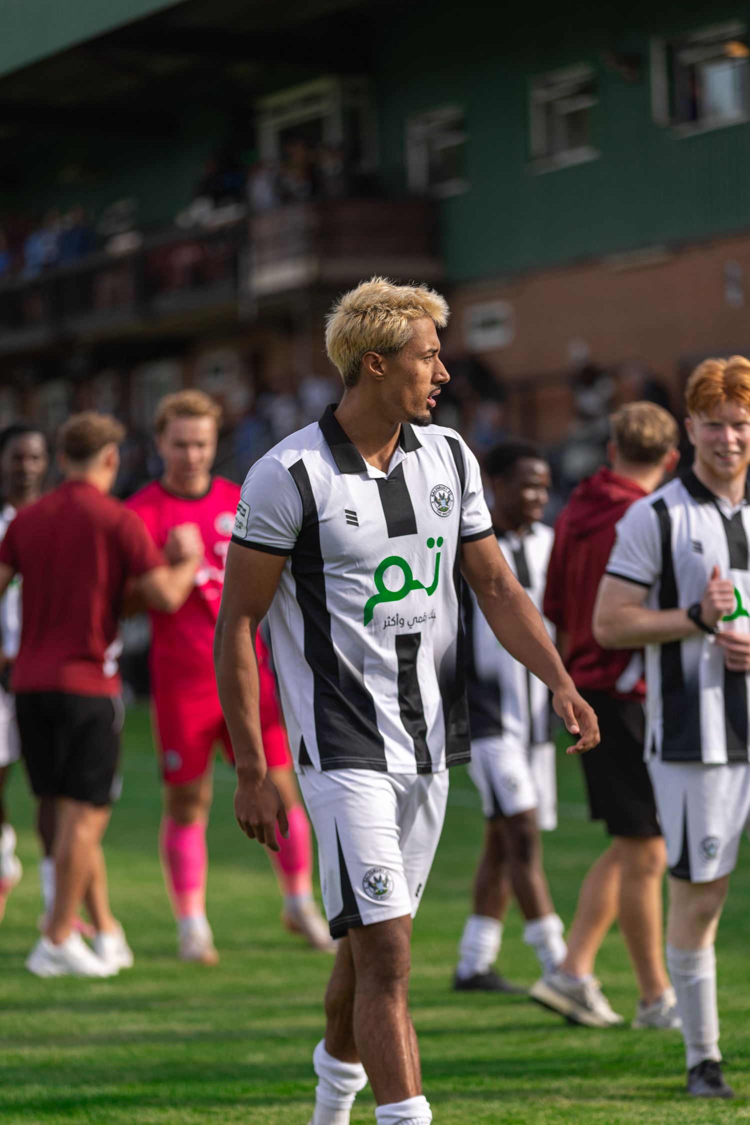 Young soccer player in black and white striped kit with blonde hair walking on pitch during a match.