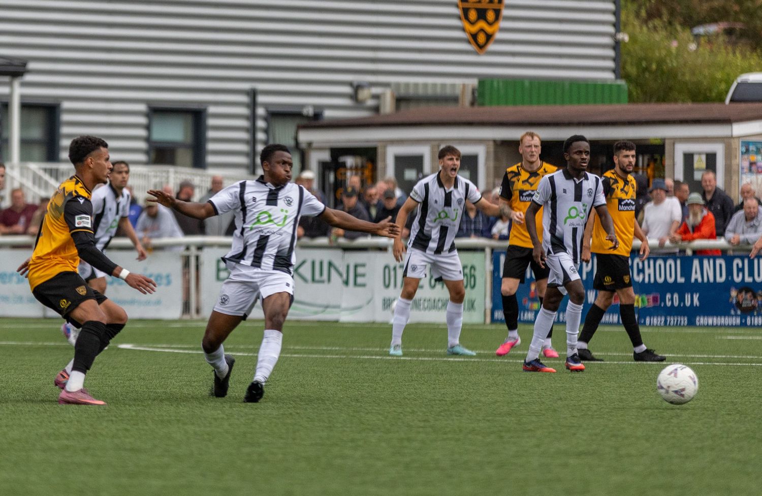 Football players in black and white vs yellow kits compete during an outdoor match on a sunny day.