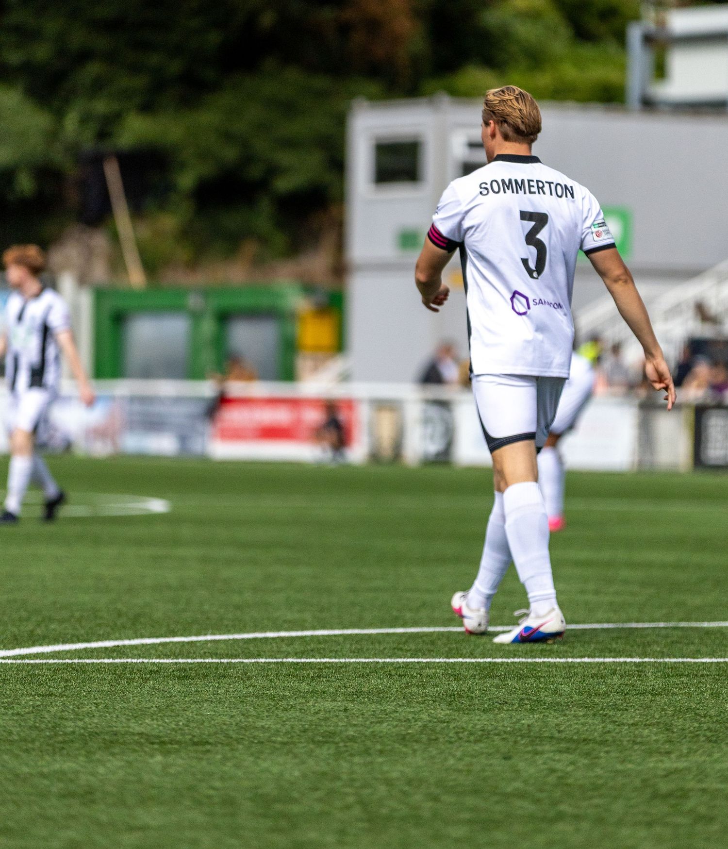 Soccer player Sommerton wearing number 3 white jersey walks on green pitch during a sunny outdoor match.