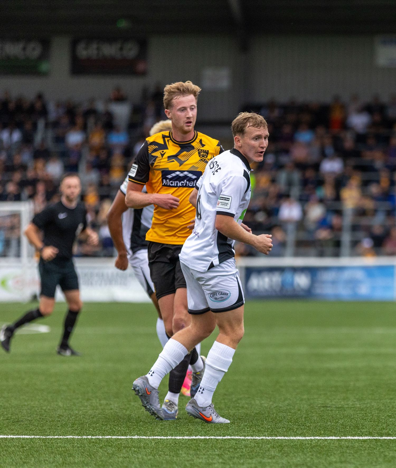 Two soccer players compete on the field during a match, with spectators and advertising boards visible in the background.