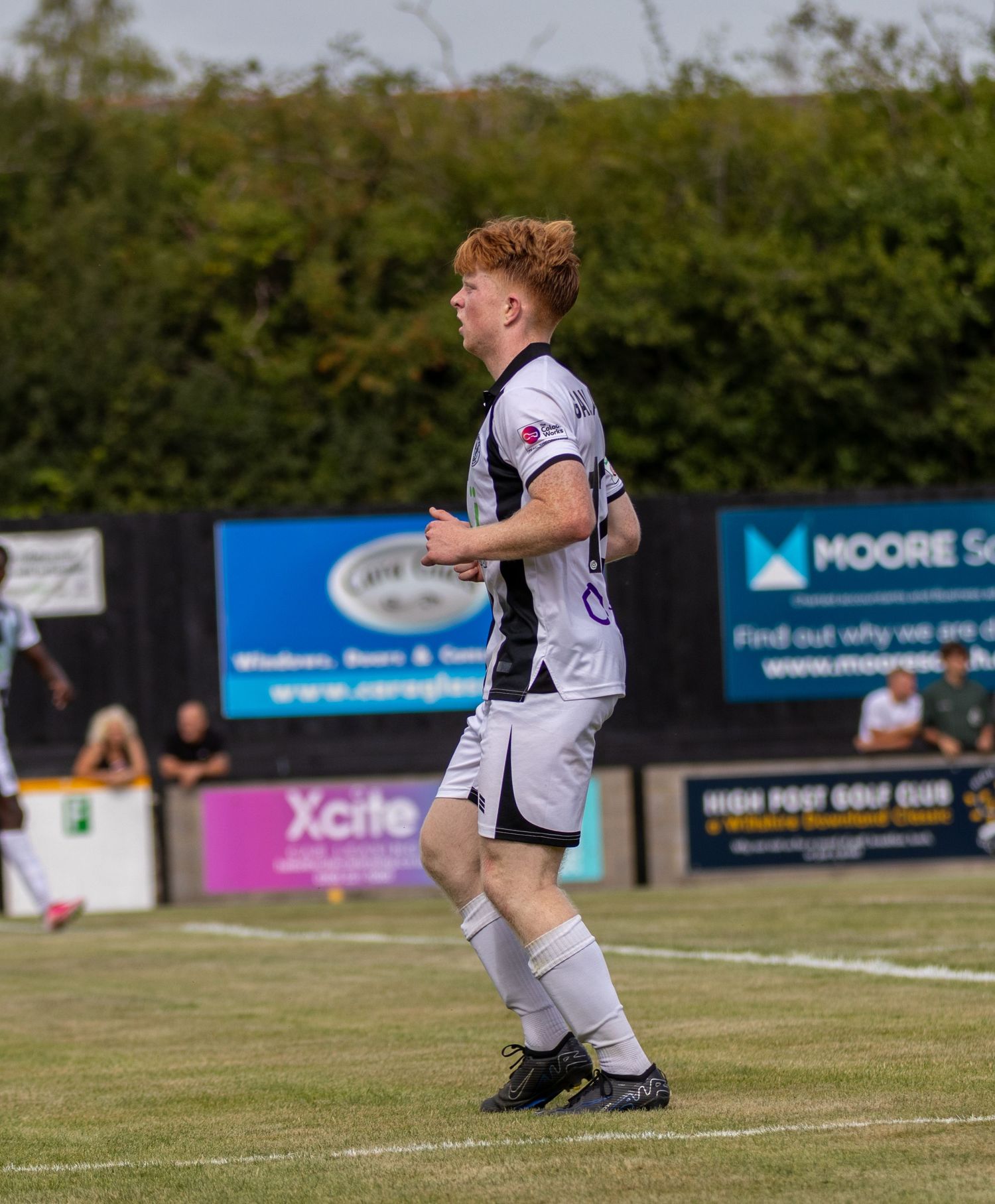 A young red-haired footballer in black and white kit jogging during a non-league football match on a sunny day.