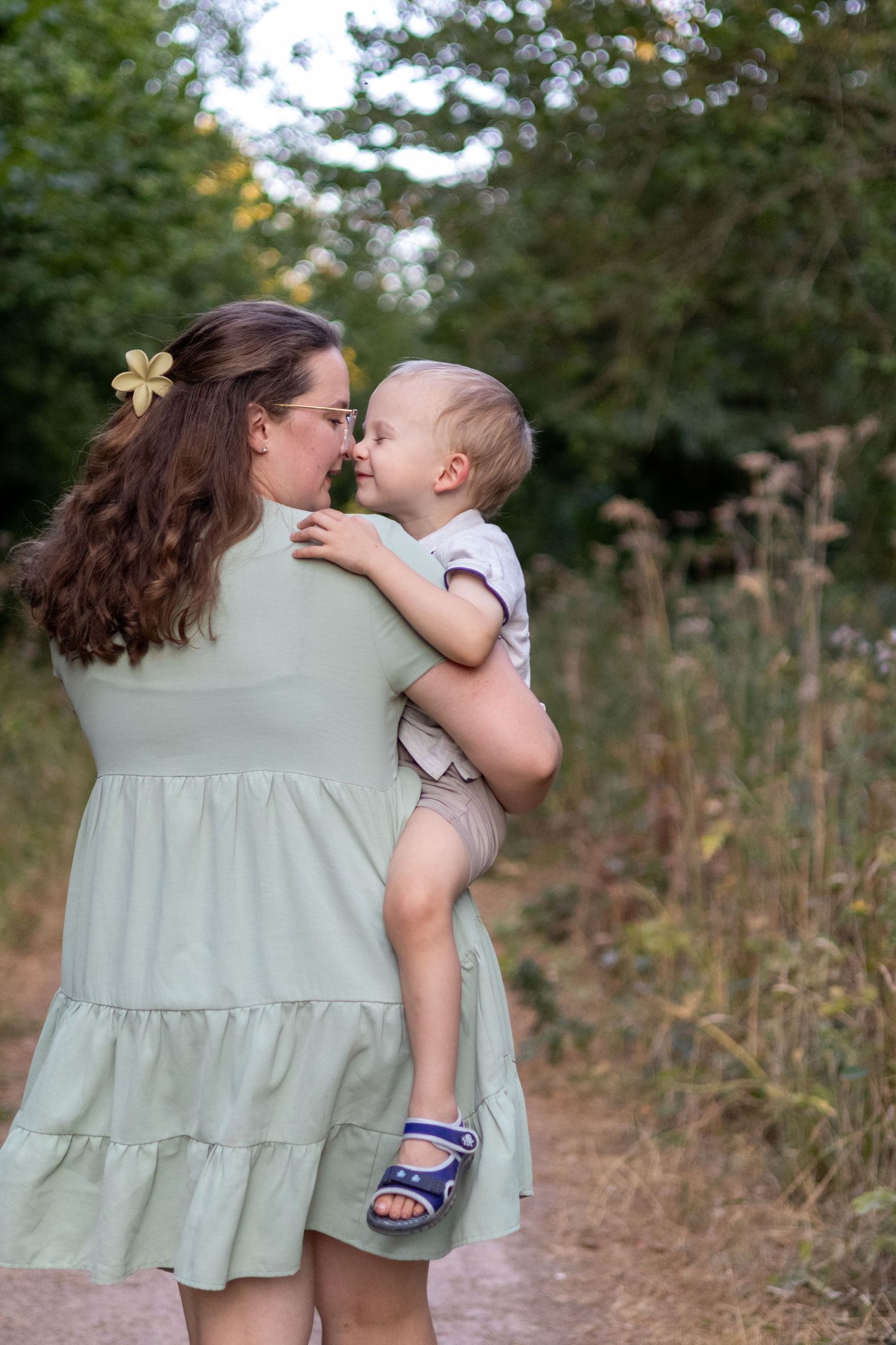 A mother in a sage green dress touches noses with her baby while standing on a peaceful nature trail.