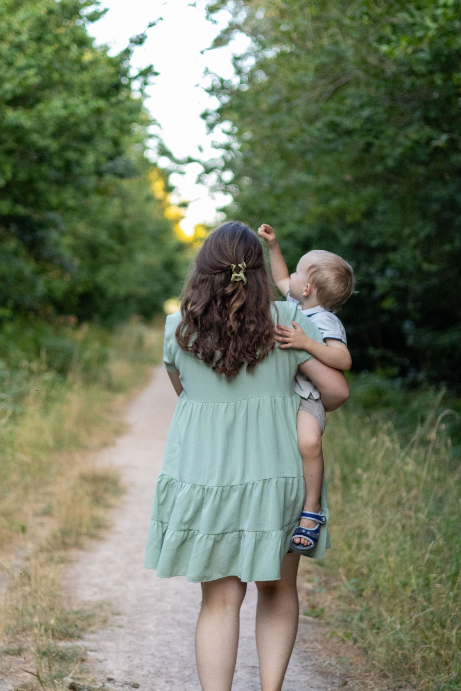 A woman in a mint green tiered dress holds her baby while walking on a forest trail.