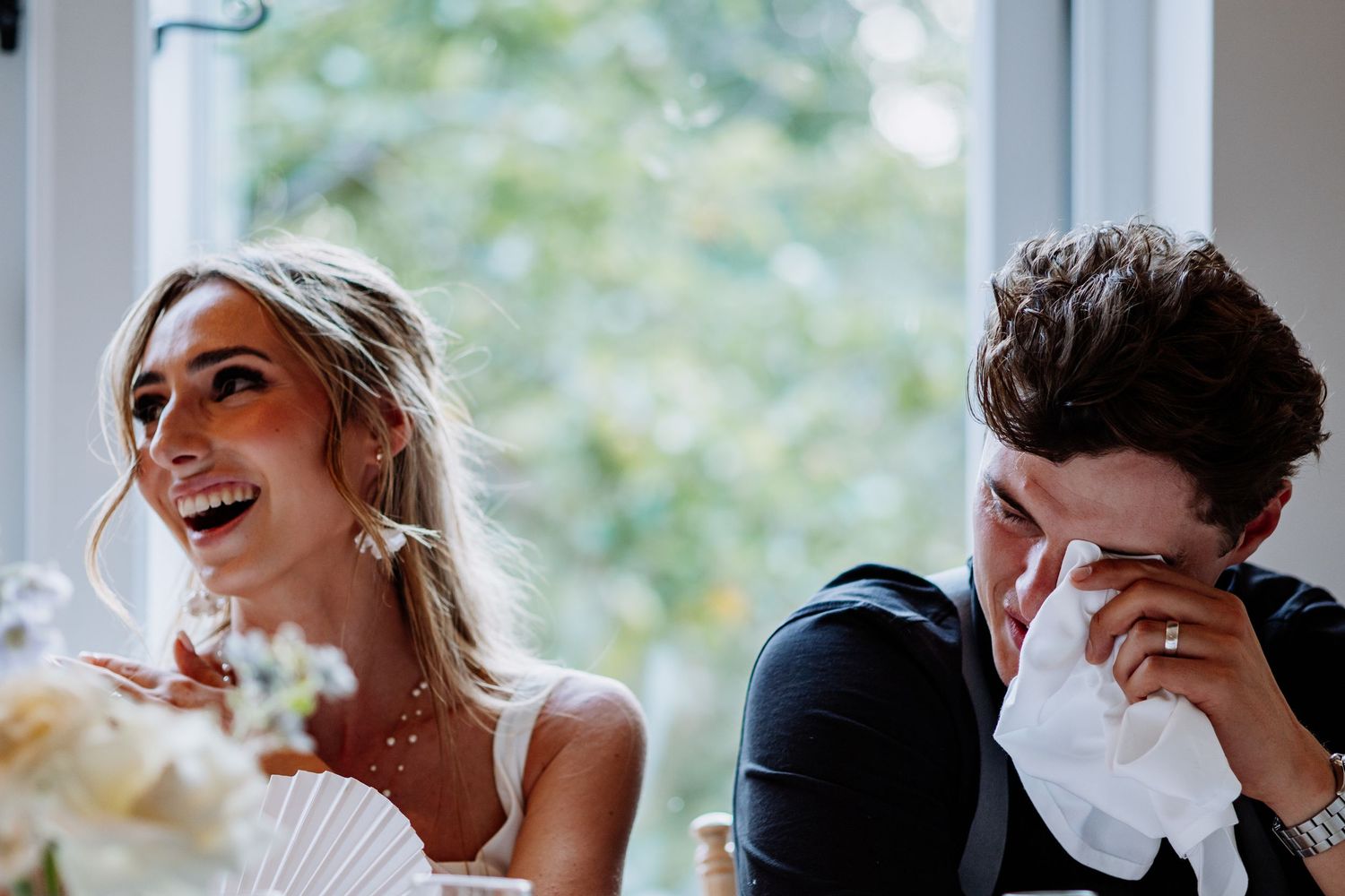 Bride laughing as emotional groom dabs eyes with tissue at wedding reception.