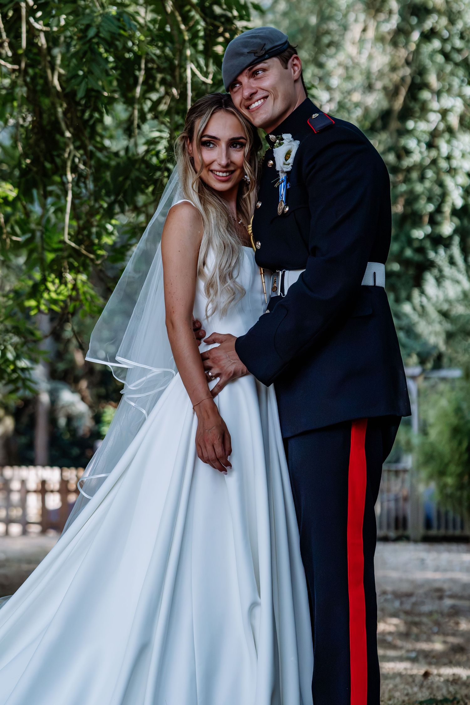 Bride in white ballgown and groom in military uniform embrace outdoors.