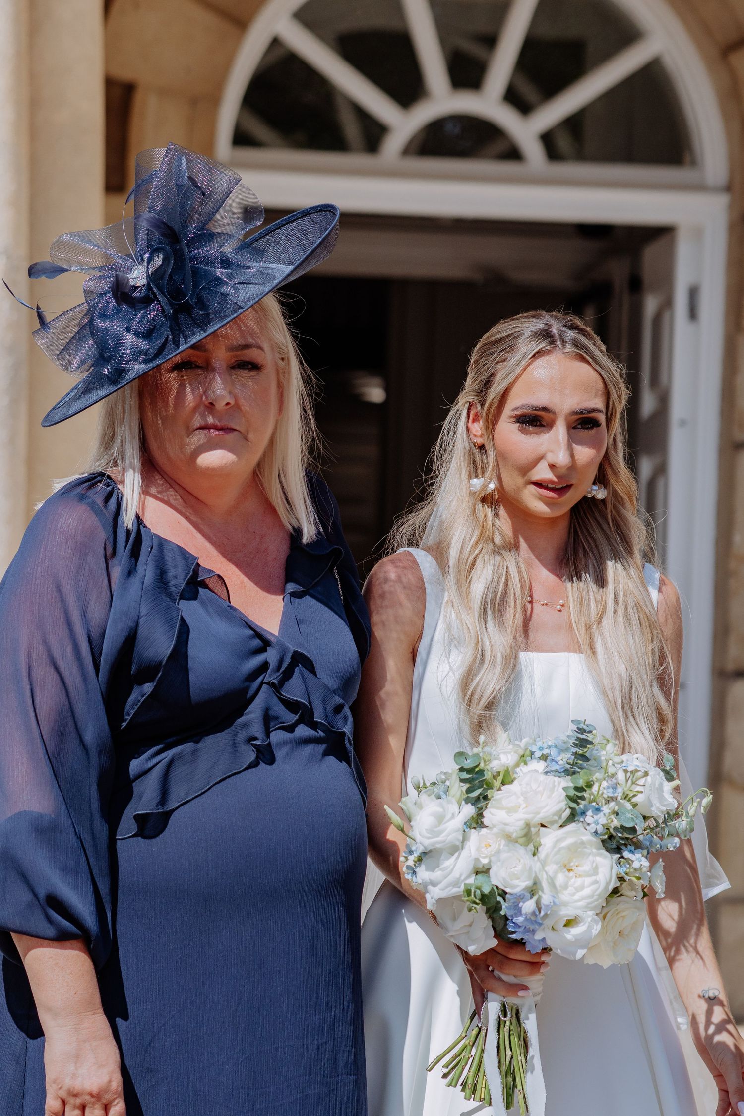Bride in white gown with bouquet stands beside woman in navy blue dress and matching fascinator hat.