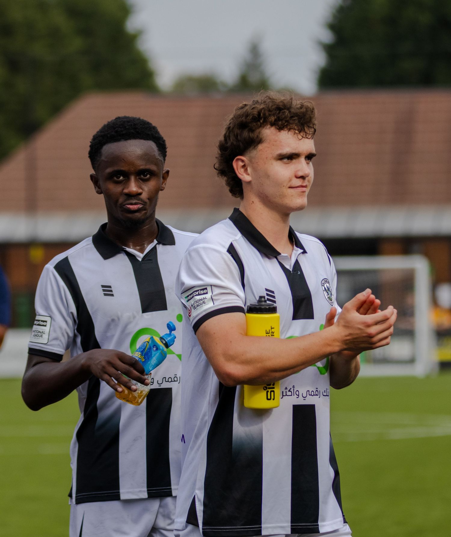 Two footballers in black and white striped kits applaud fans after a match at a football ground.