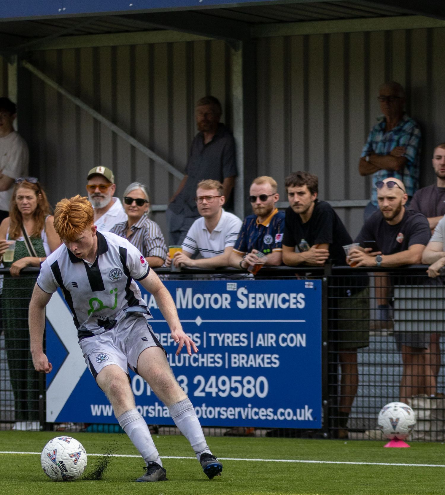 Red-haired footballer in white kit dribbles ball during match at Horsham FC ground with spectators watching.