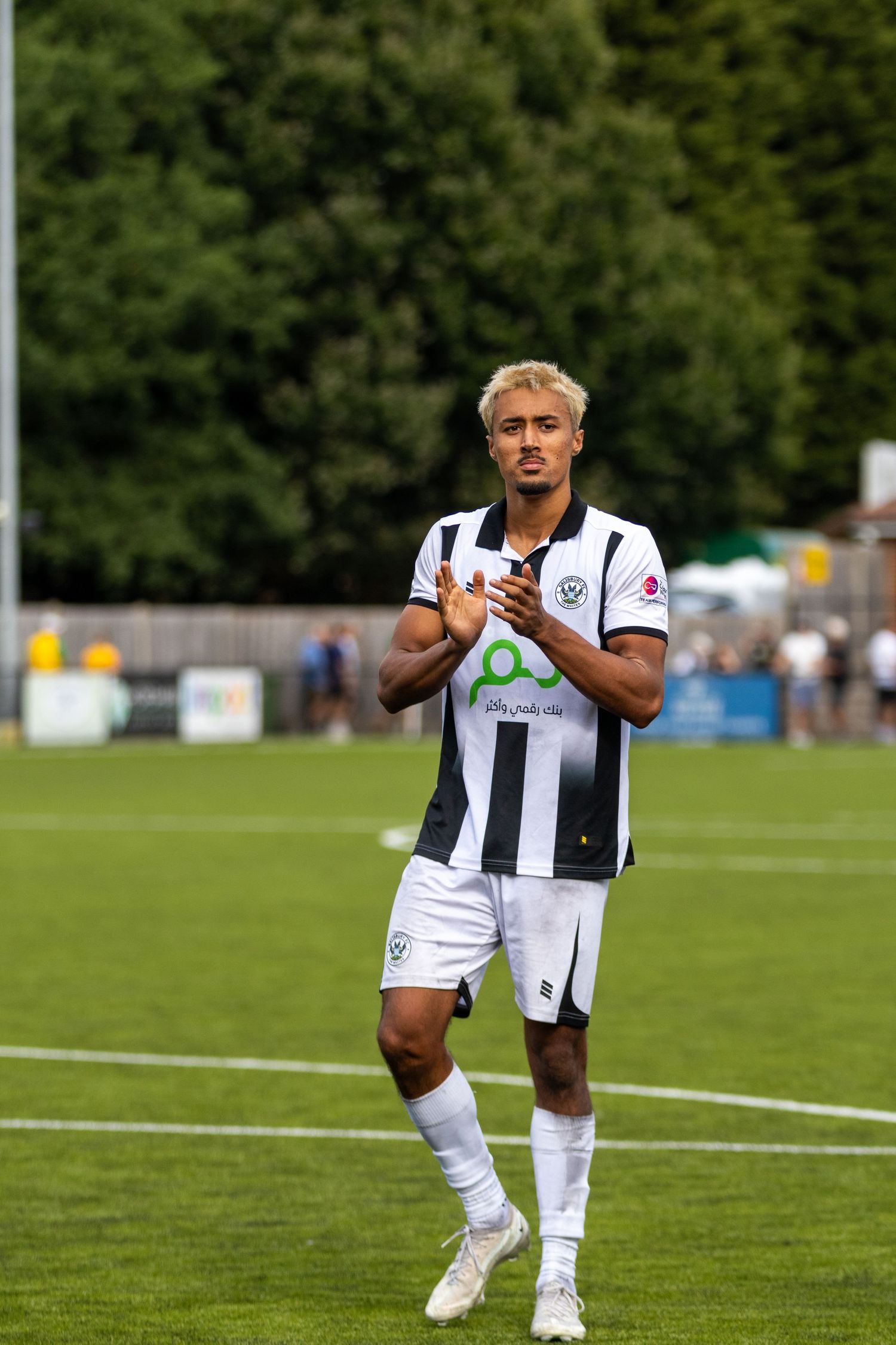 A footballer in black and white striped kit applauds fans on a green pitch during a sunny match day.