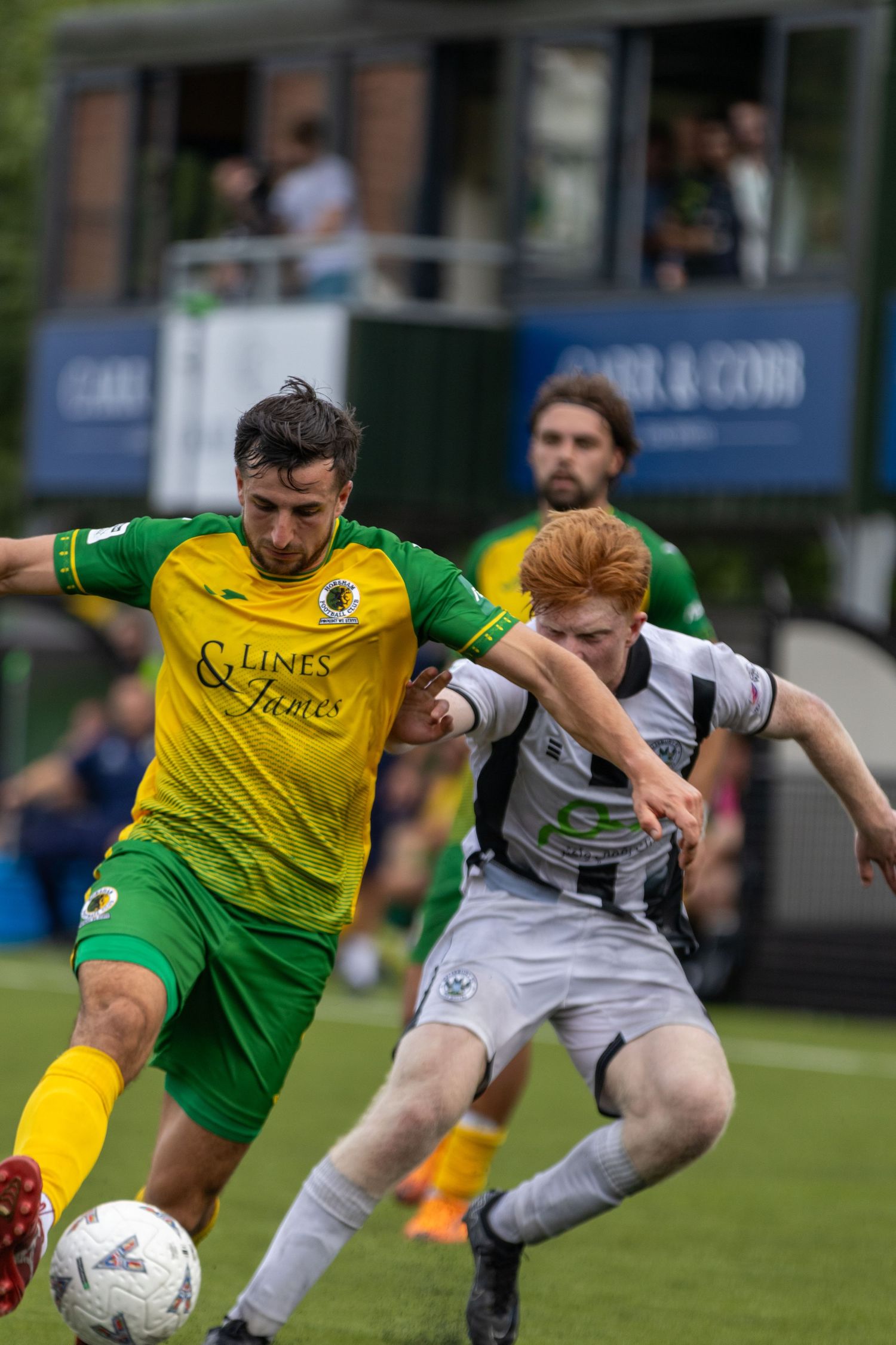 Red-haired player in white kit battles for ball against yellow-green opponent during competitive football match.
