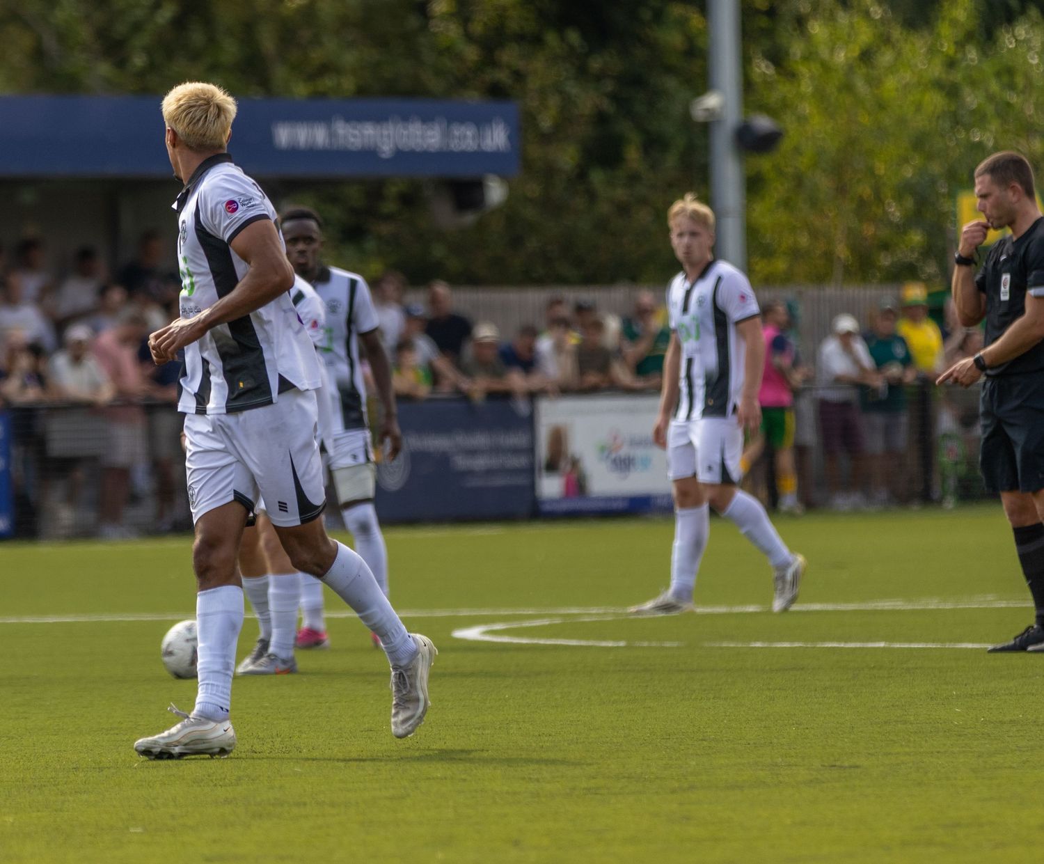 Footballers in black and white striped kits prepare for a set piece on a sunny green pitch during a competitive match.