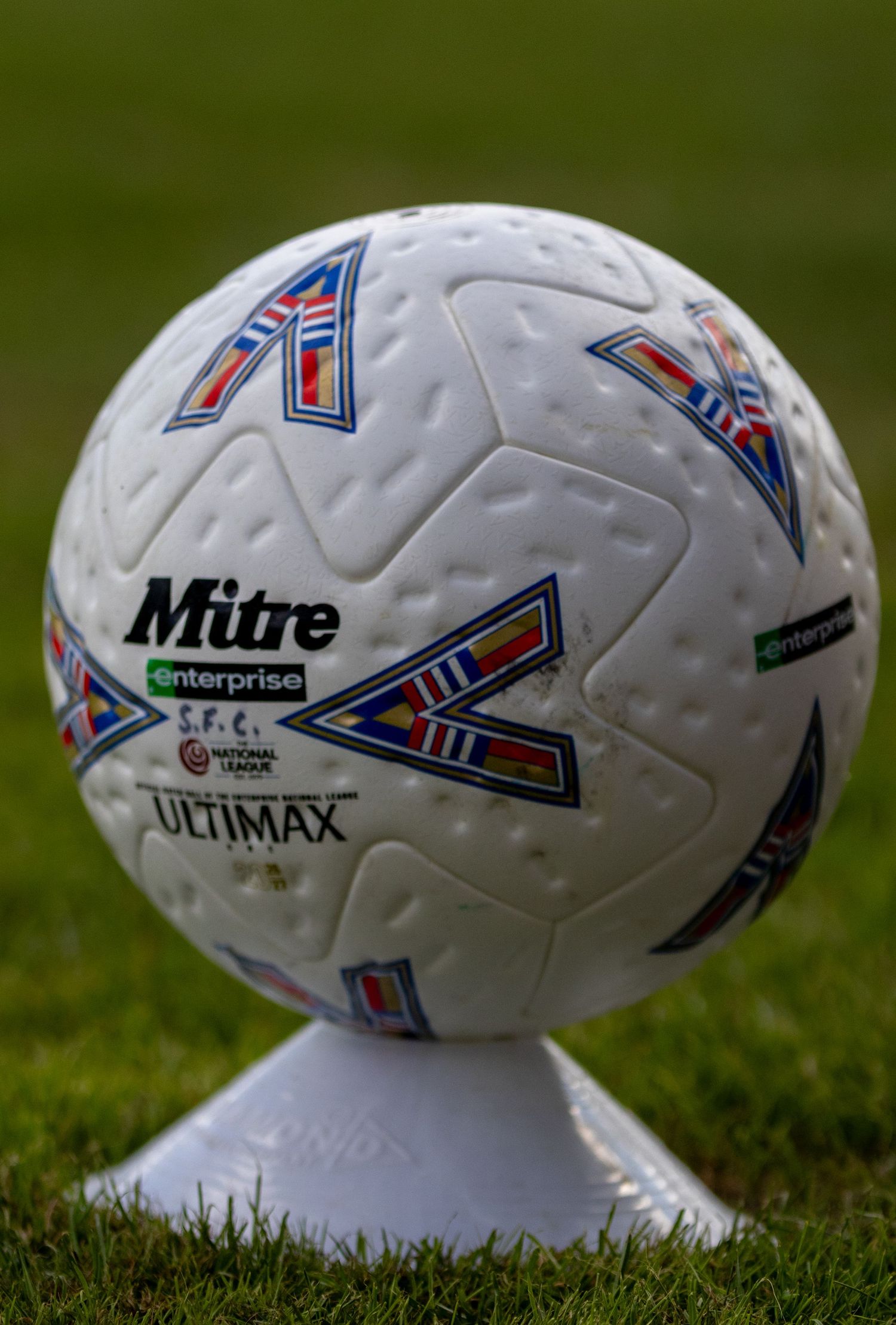 Mitre Ultimax National League football on white tee displayed on green grass pitch.