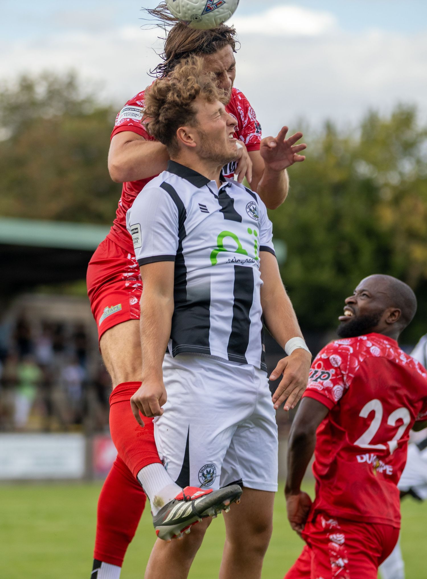 Two soccer players contest a header during a match, with red and black-and-white kits visible on a sunny day.
