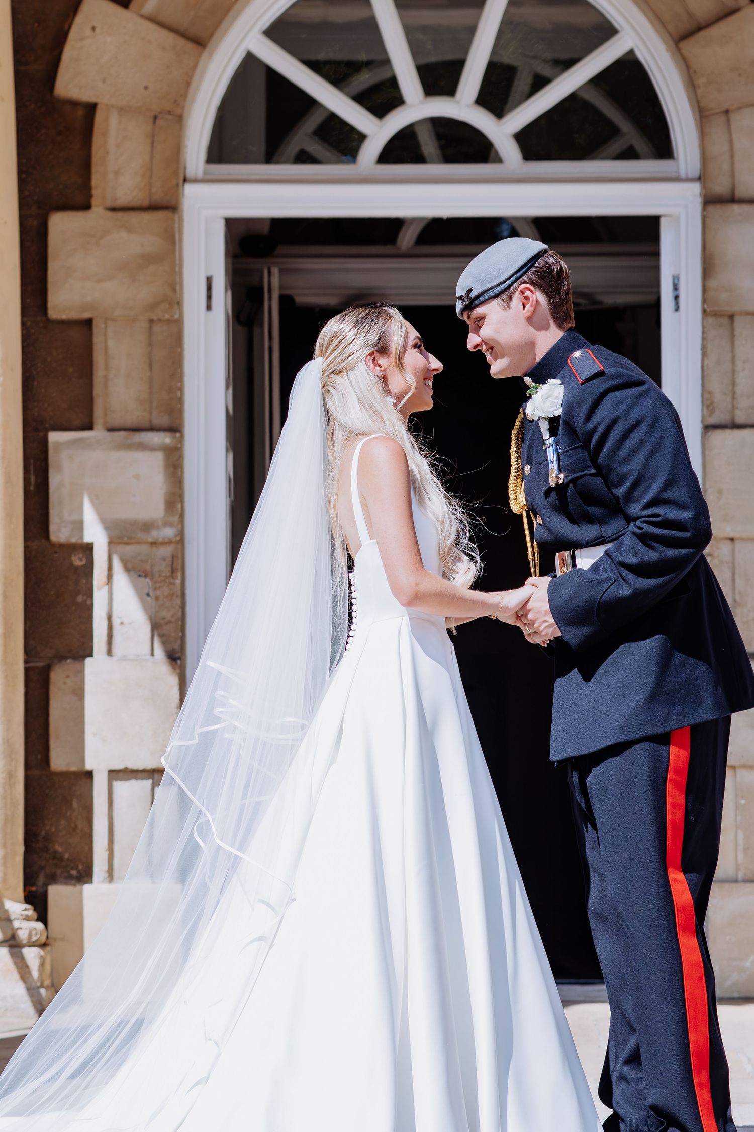 Bride in white gown and veil holds hands with groom in military uniform outside a grand stone building.