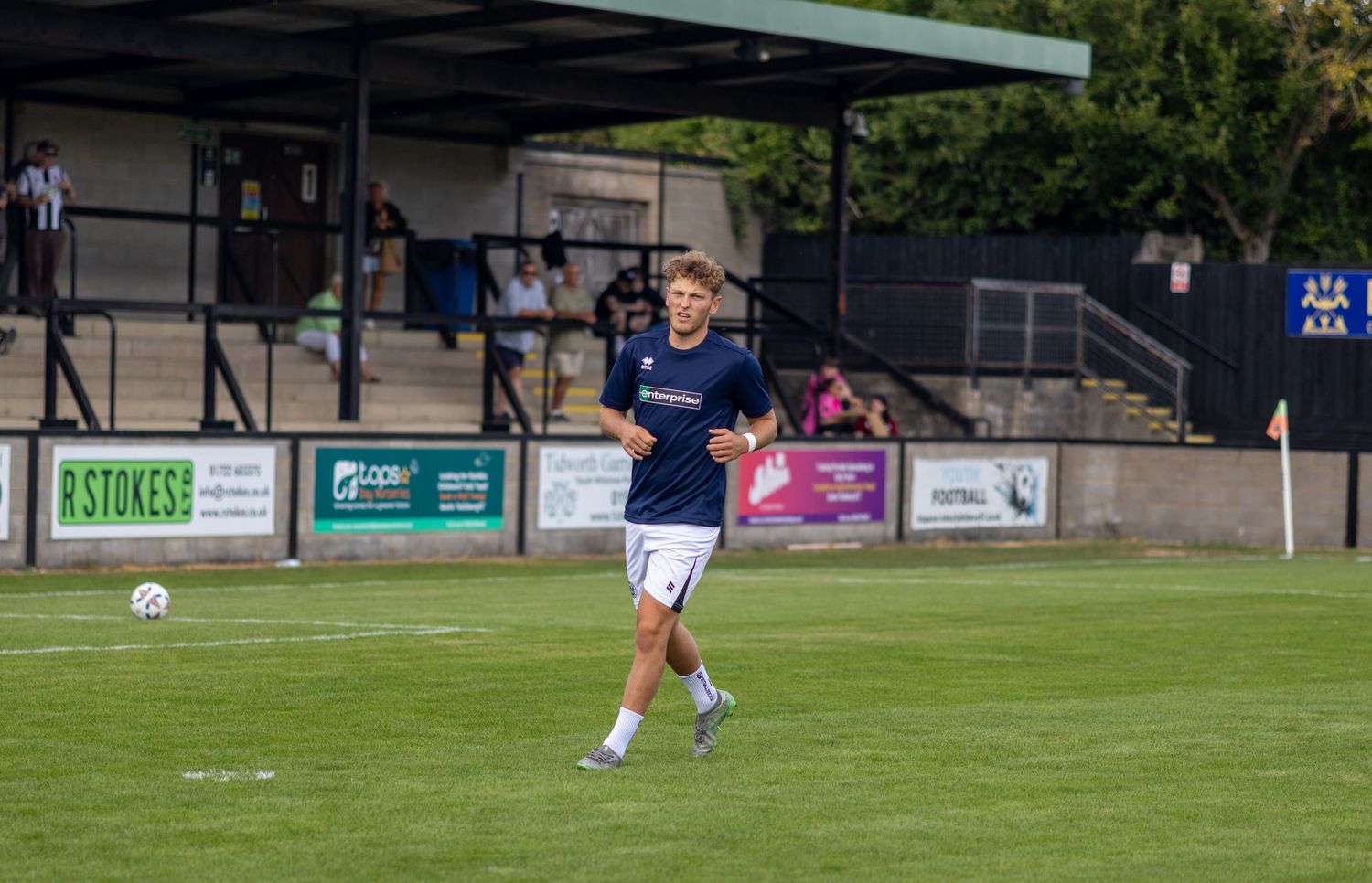 Male footballer in navy blue uniform running with the ball during warmup at a non-league football ground.