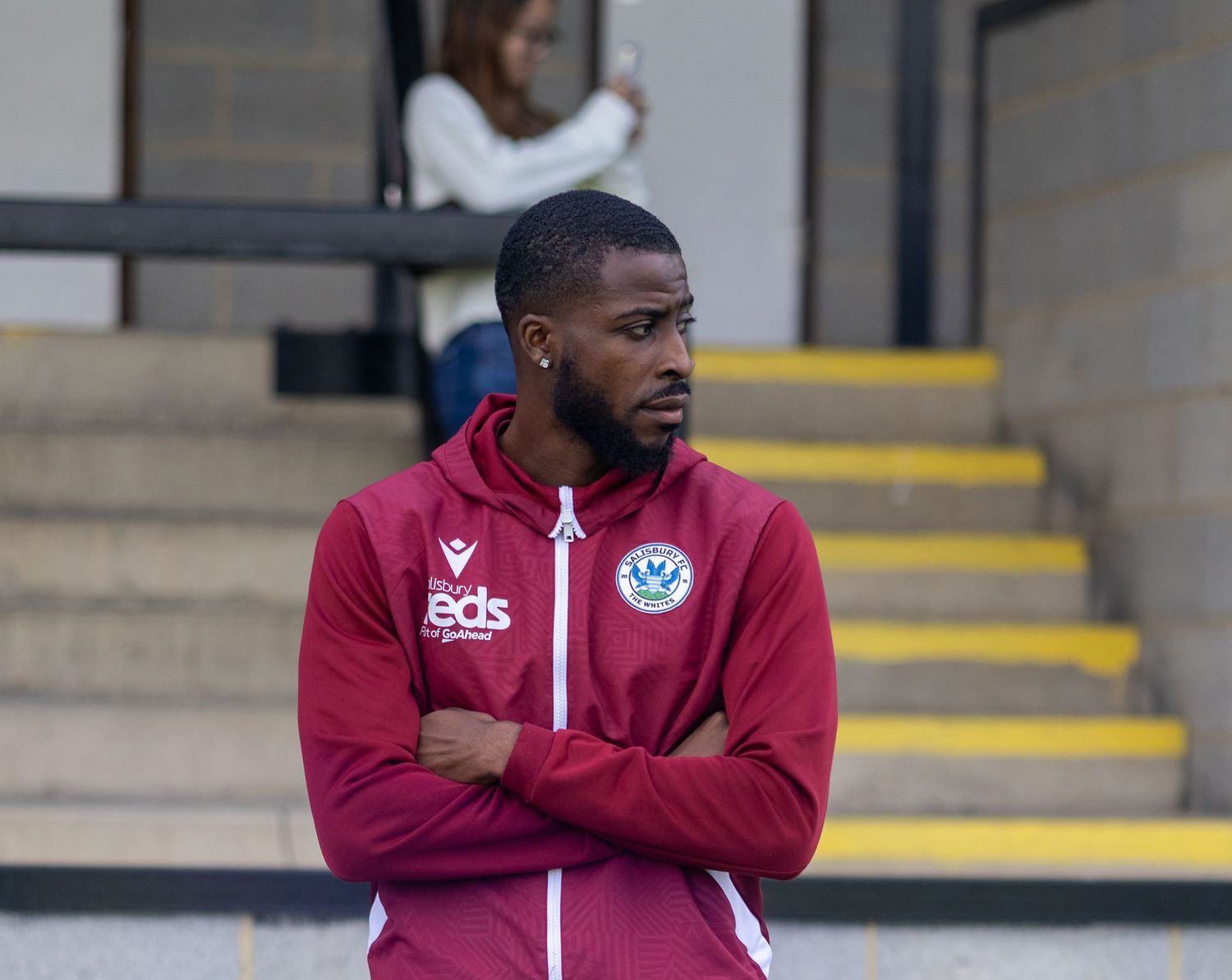 Man in maroon sports jacket with arms crossed standing near yellow-edged steps outdoors.