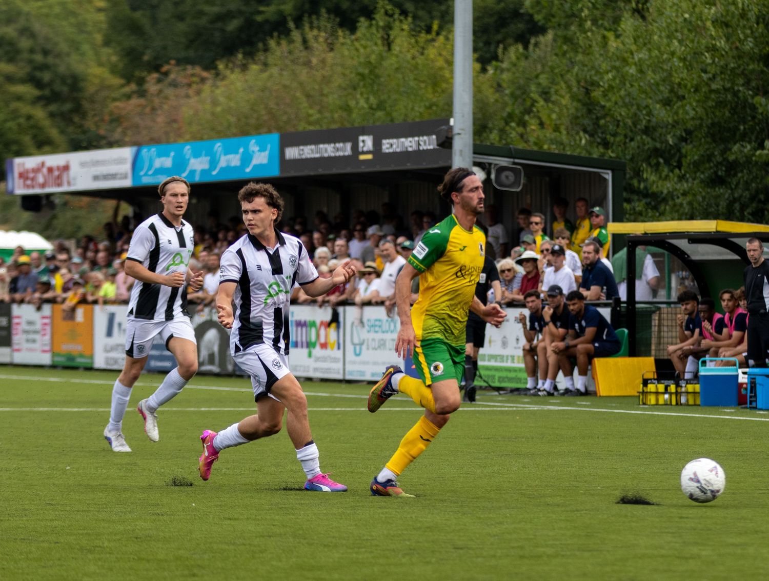 A footballer in yellow kit sprints with the ball as black and white opponents give chase during a sunny outdoor match.