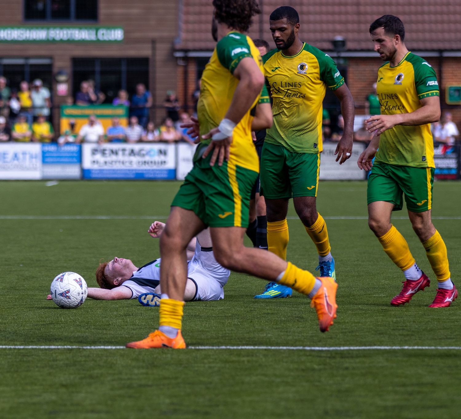 White kit player on ground with ball surrounded by three yellow and green opponents on pitch.