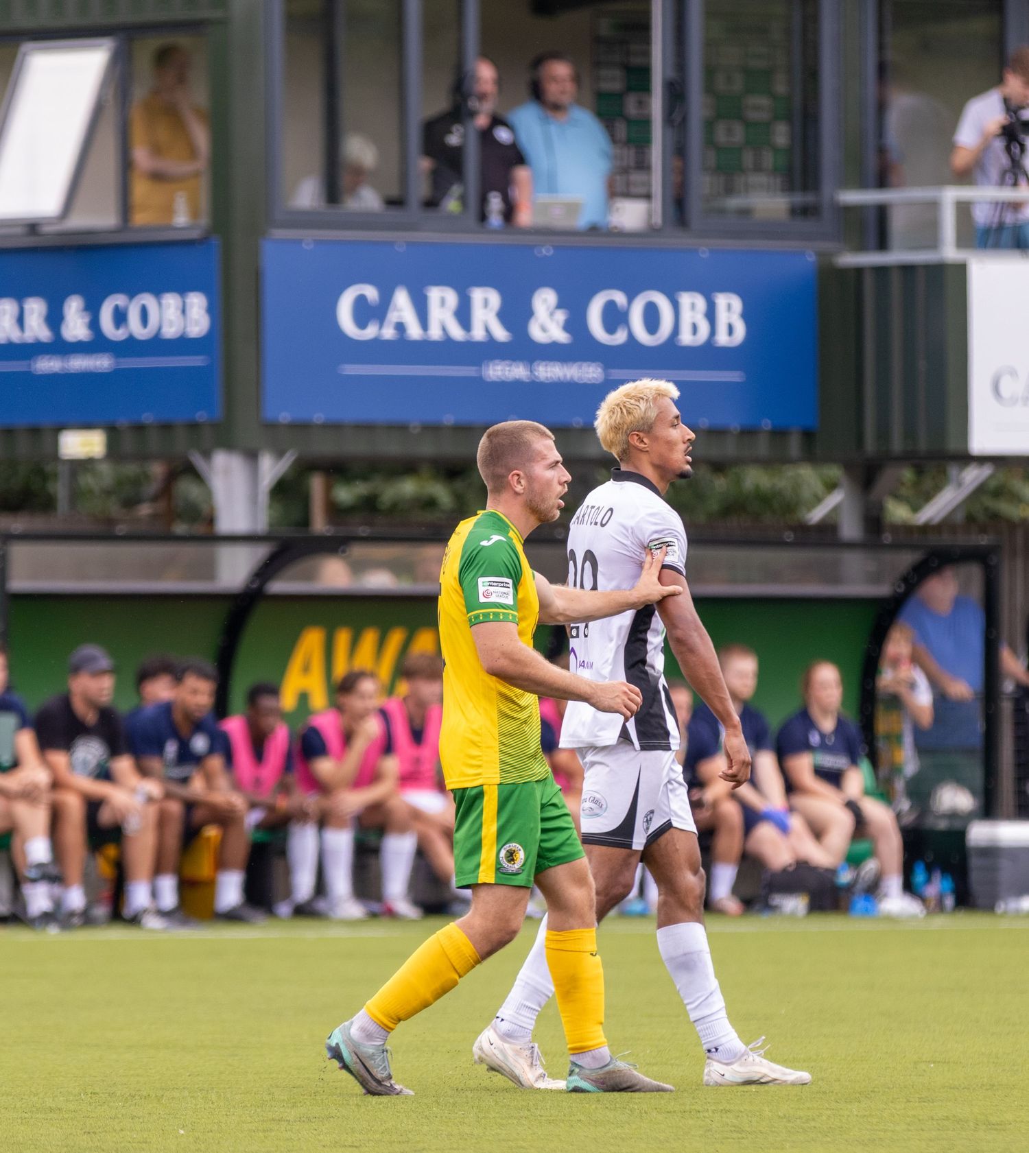 Two footballers, one in yellow-green and one in white, exchange words near the Carr and Cobb stand.