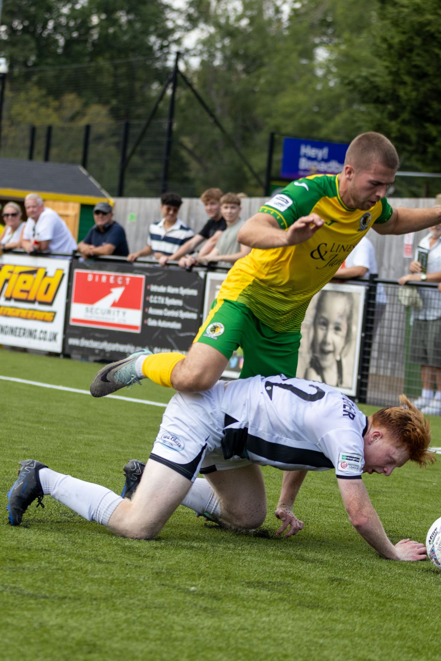 Two footballers challenge for the ball, one in yellow-green kit and one in white, during a match.