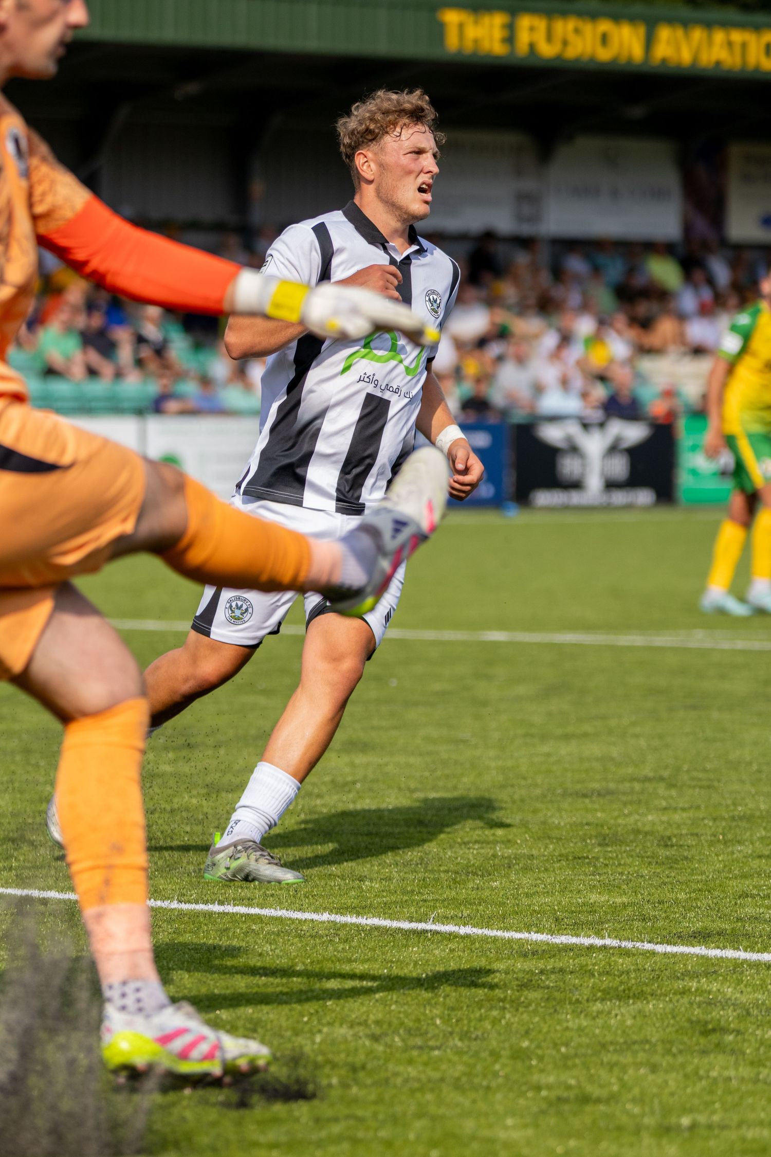 A footballer in black and white striped kit attempts a shot as goalkeeper approaches during outdoor match.
