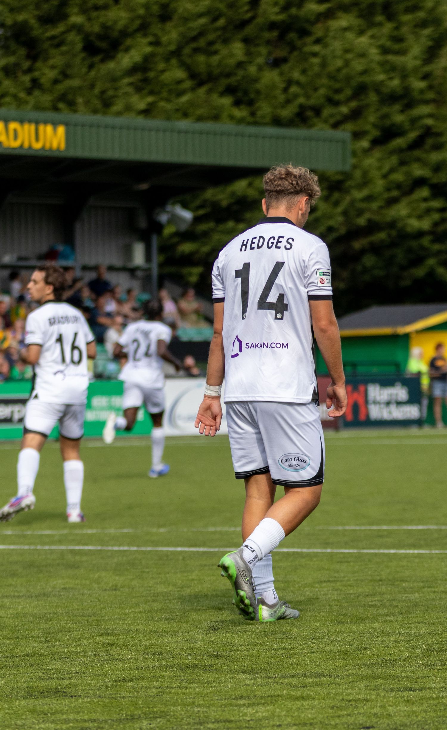 Footballer Hedges #14 in white Sakin.com sponsored kit walks across the pitch during a non-league football match.