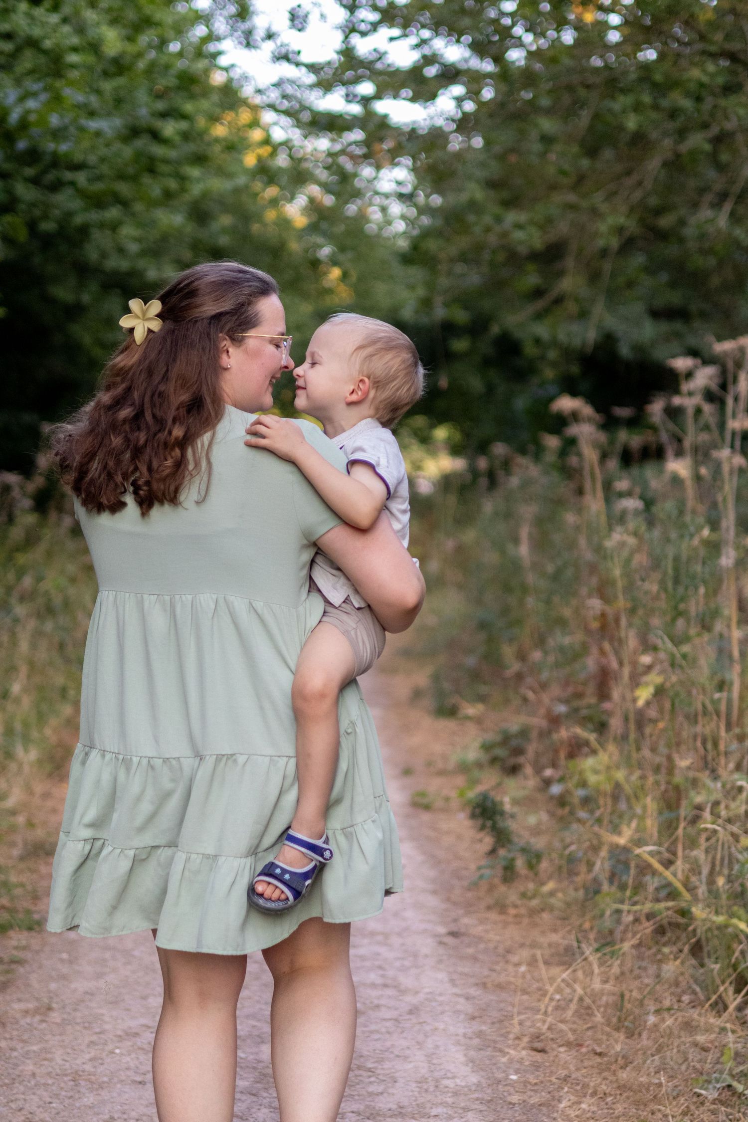 A mother with a flower hair clip holds her laughing baby close on a scenic outdoor nature trail.