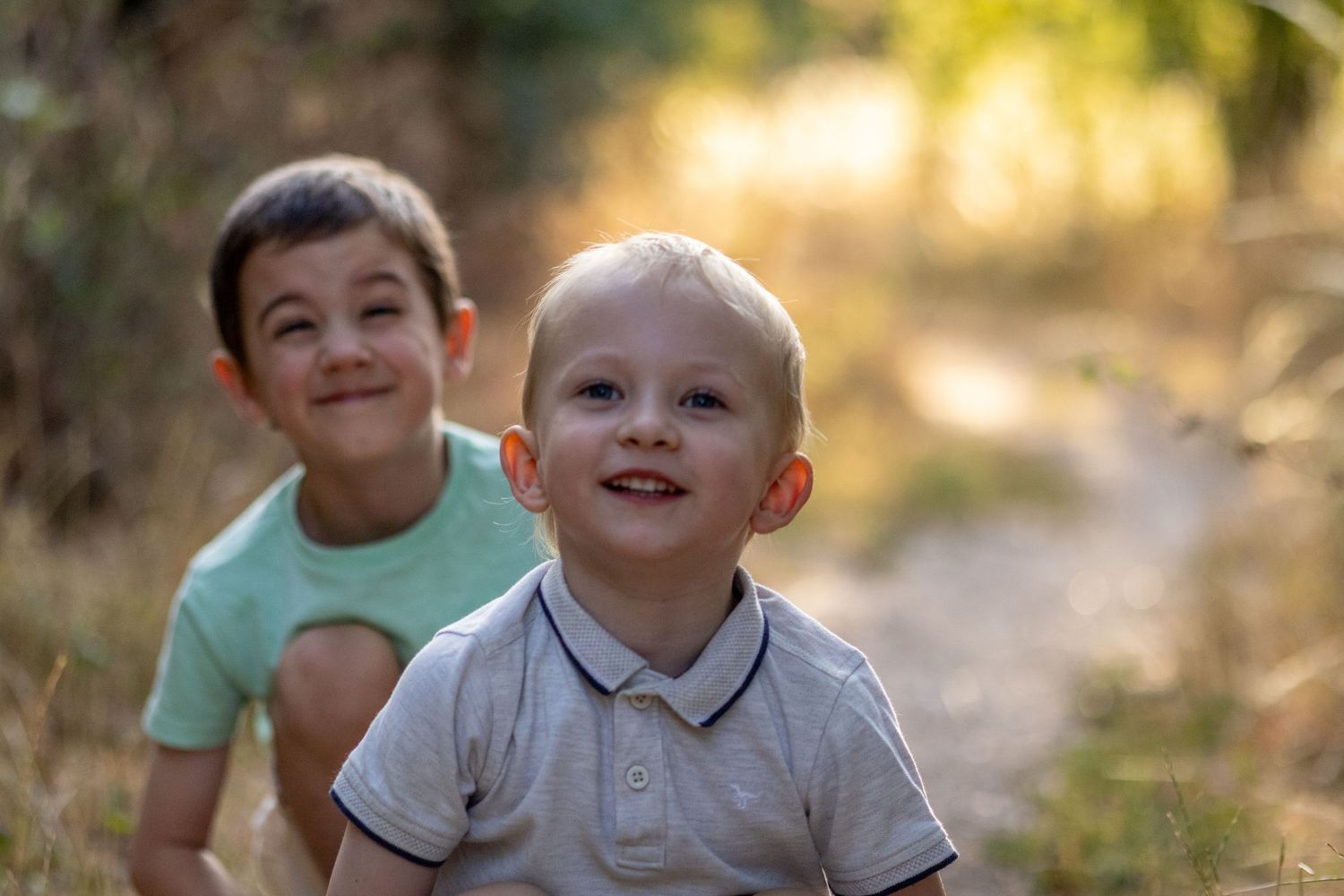 Two smiling young boys sitting together on a sunlit dirt path in a natural outdoor setting.