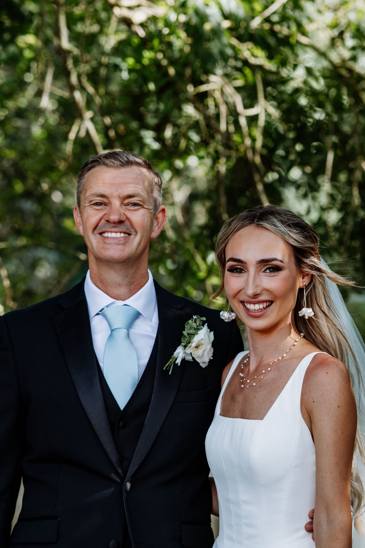 Wedding couple posing outdoors, groom in black suit with light blue tie, bride in elegant white dress.