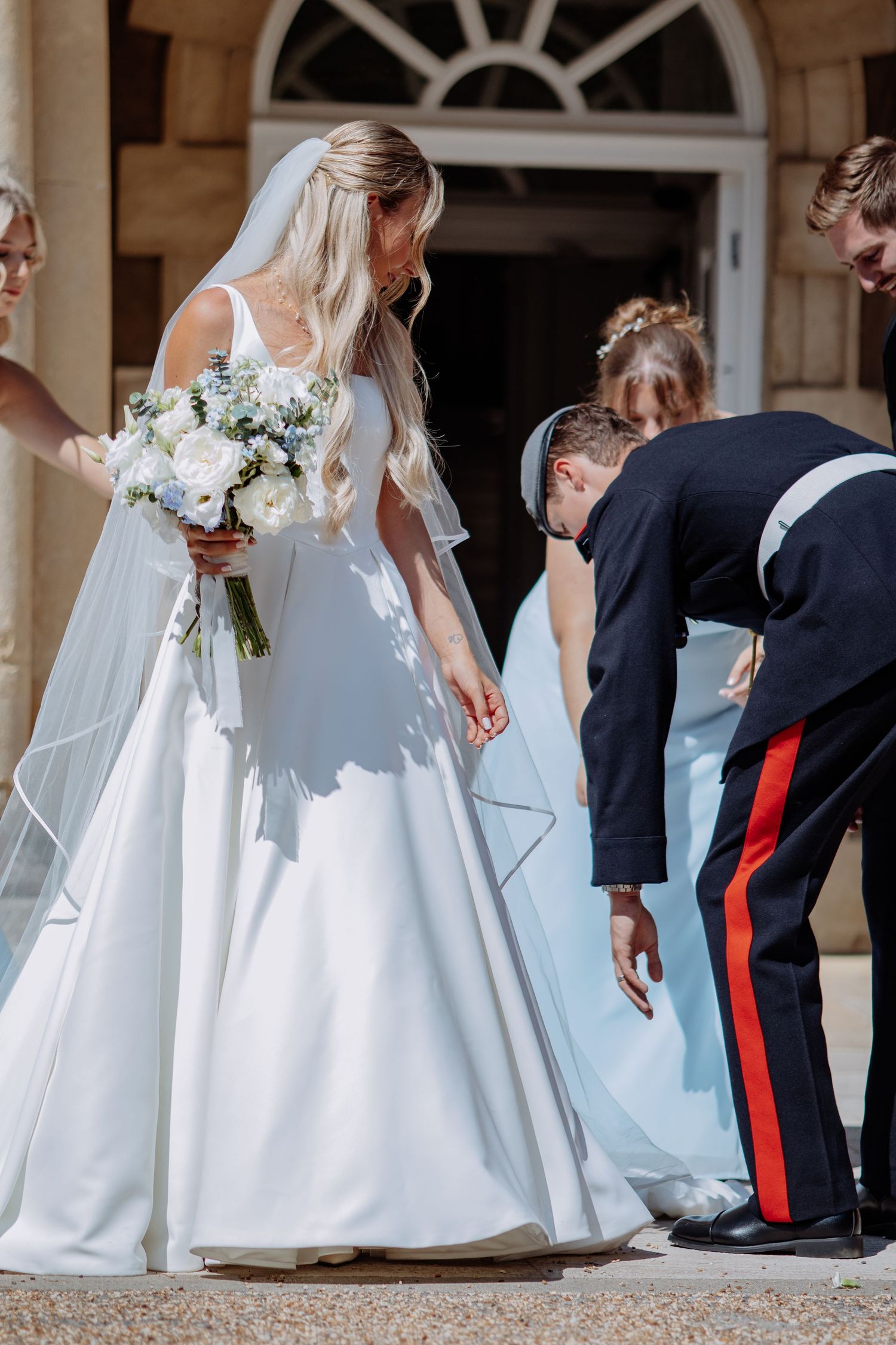 Military groom adjusts bride's dress train outside grand wedding venue.