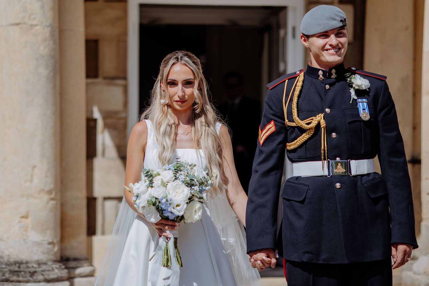 Newlywed couple walks hand in hand outdoors, bride in white ballgown with blue bouquet and groom in military uniform.