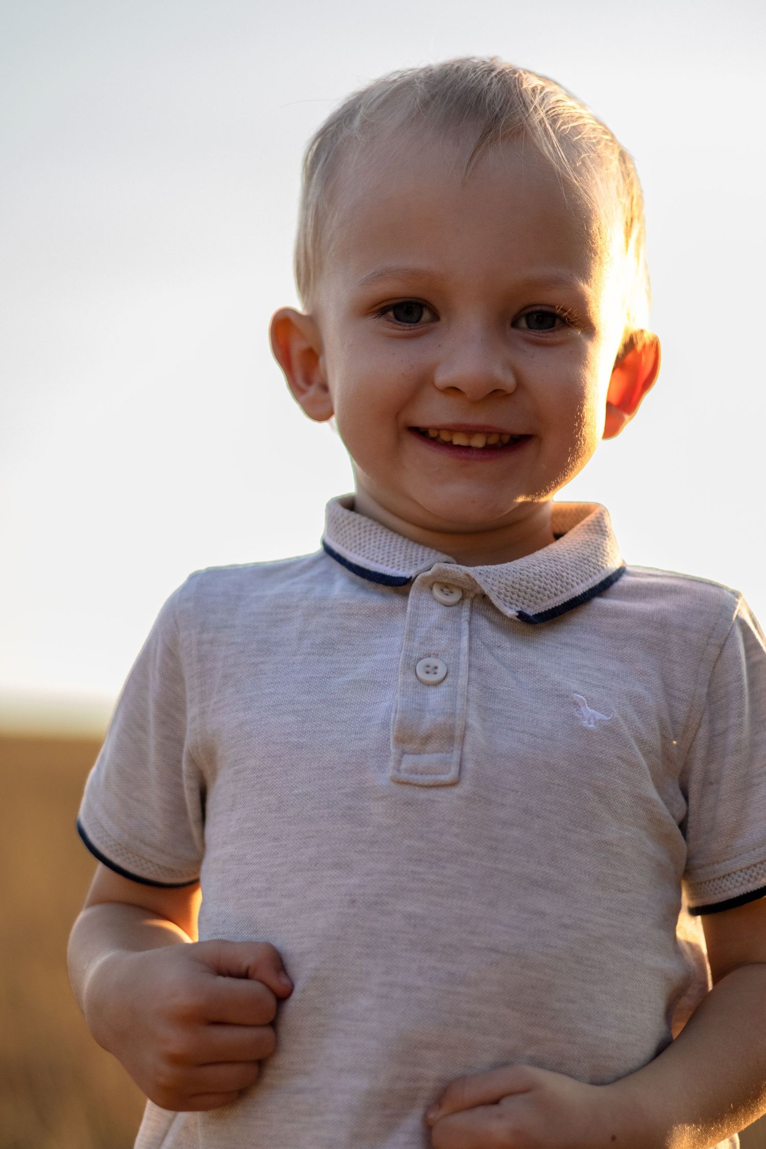 A cute smiling blond toddler boy in a gray polo shirt stands outdoors in bright sunlight.