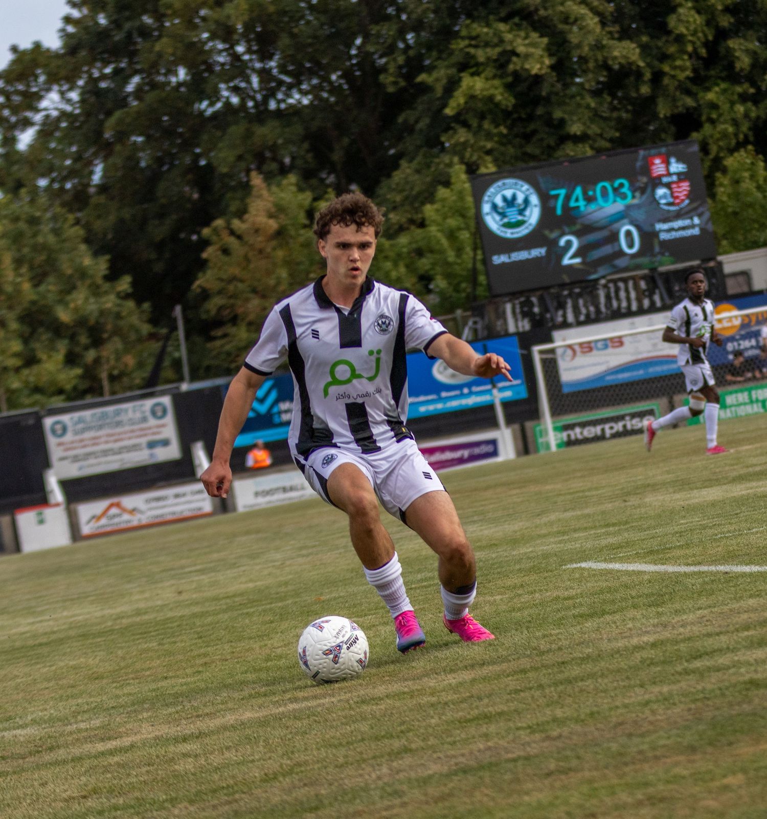 A young footballer in black and white striped kit dribbles the ball on a sunny matchday pitch.
