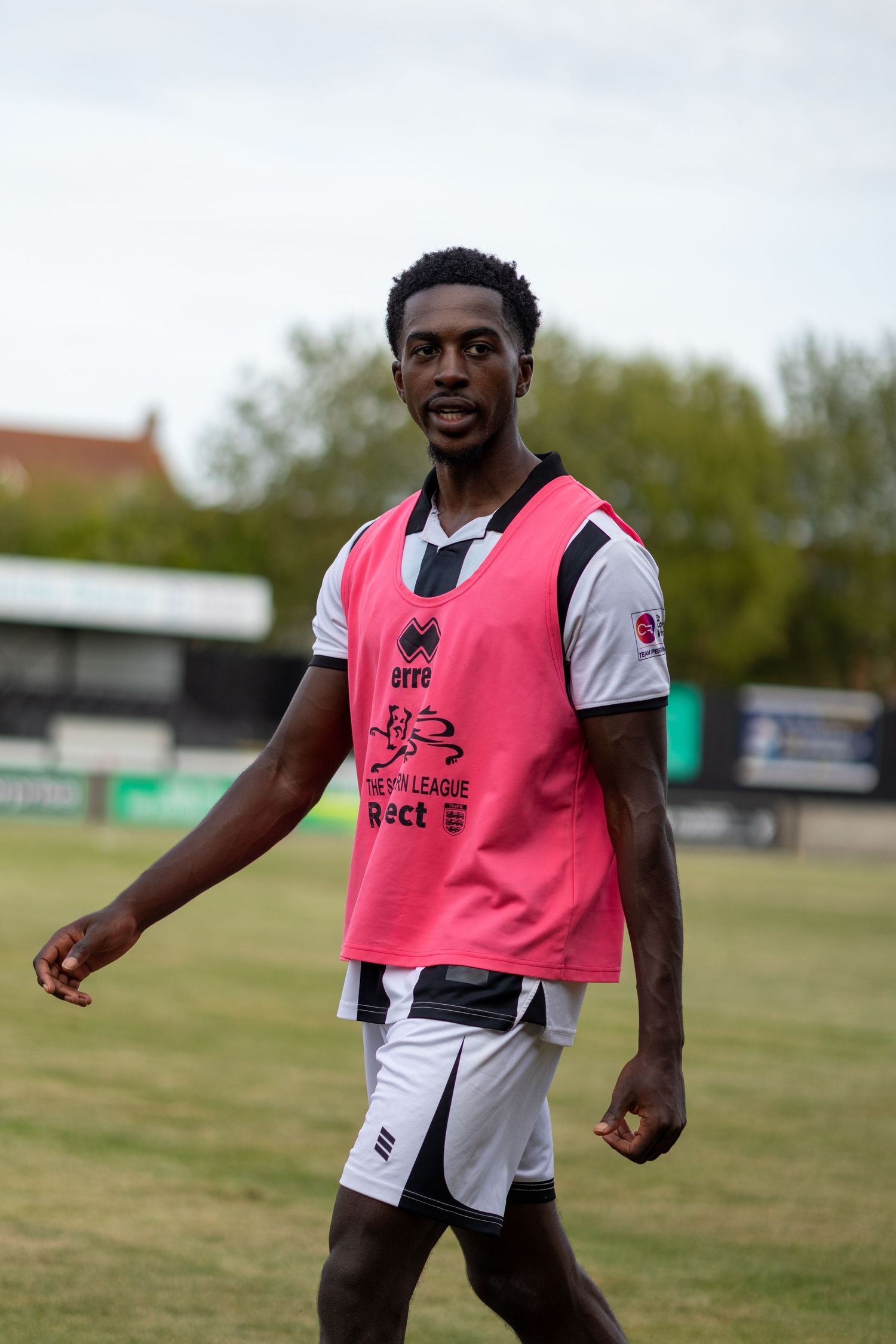 A male soccer player wearing a pink training bib over a white and black kit walks on a grass field.