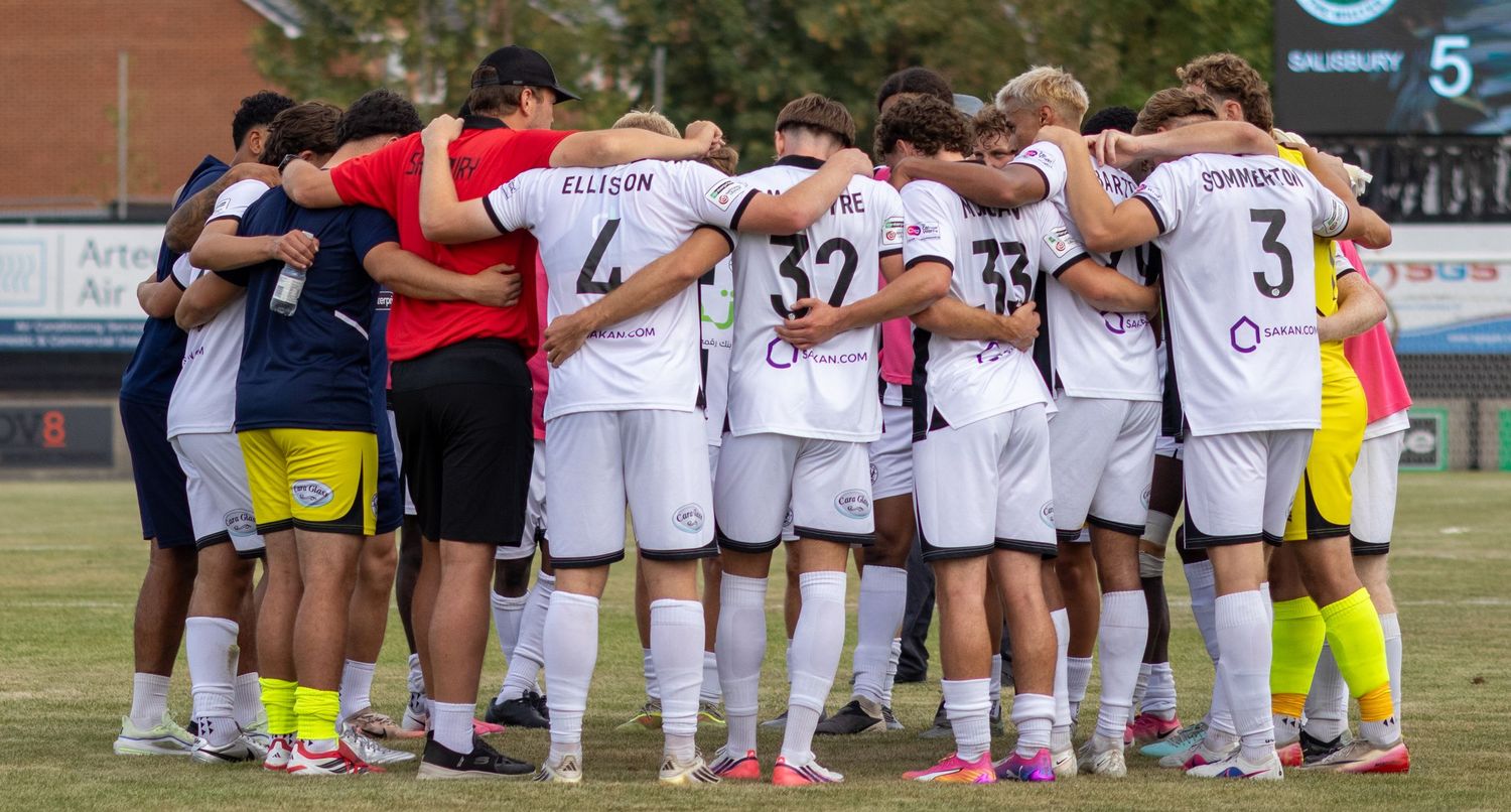 Soccer team in white uniforms forms a pre-match huddle on green field before game.