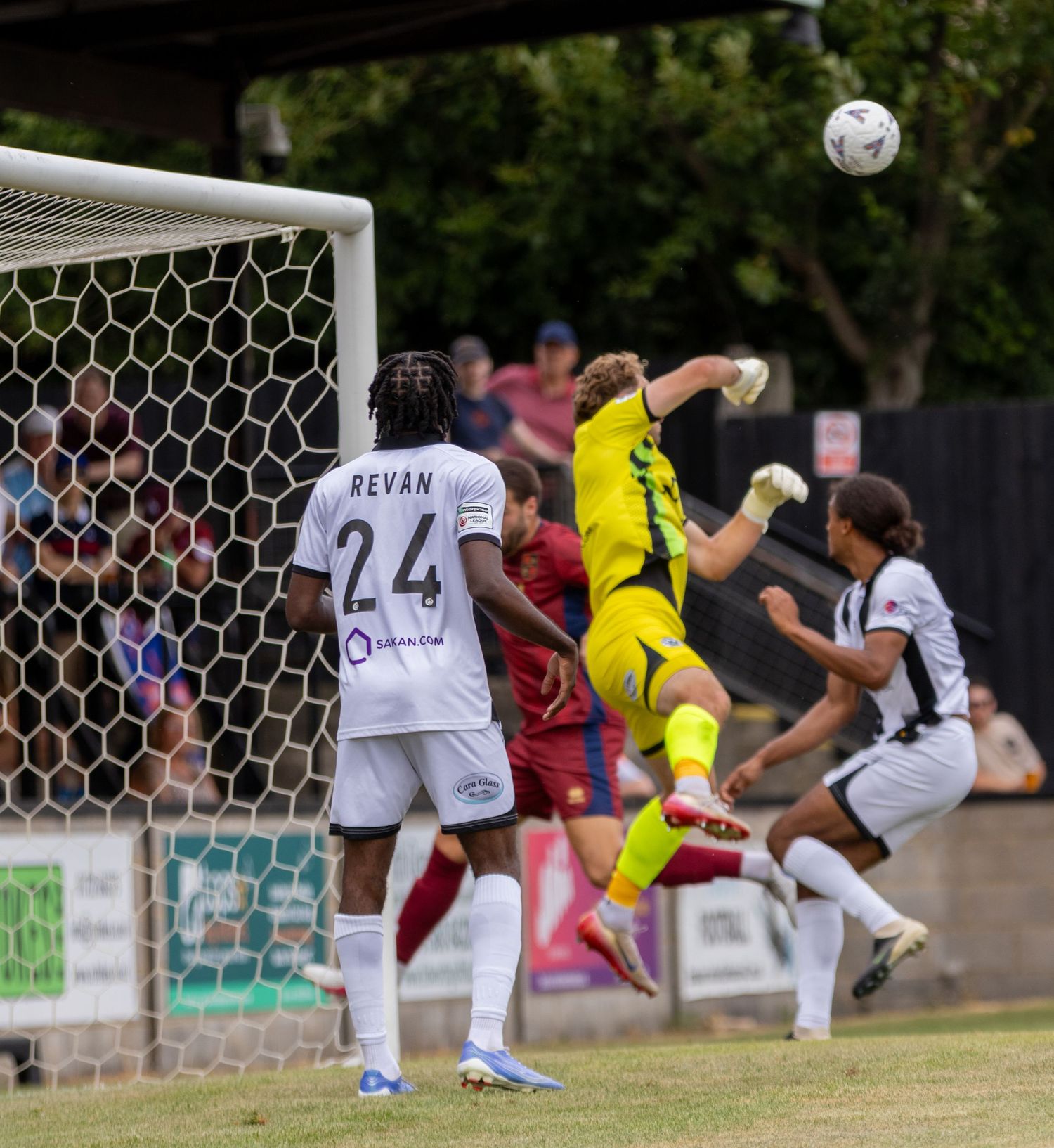 A goalkeeper in yellow kit punches the ball clear during a football match near the goal.