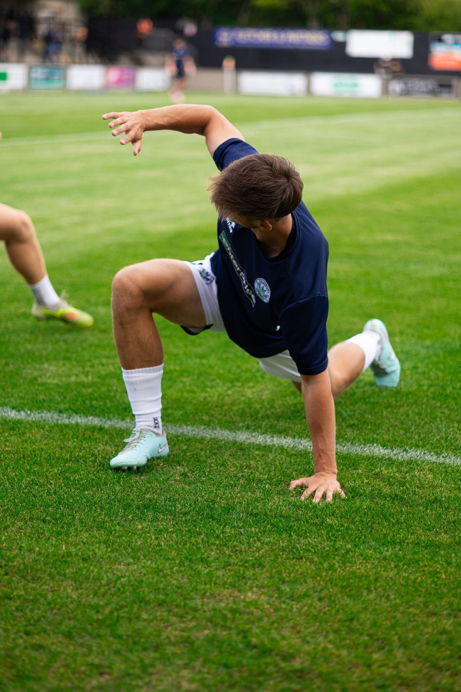 Soccer player in navy kit stretching on green grass pitch during pre-match warmup routine.