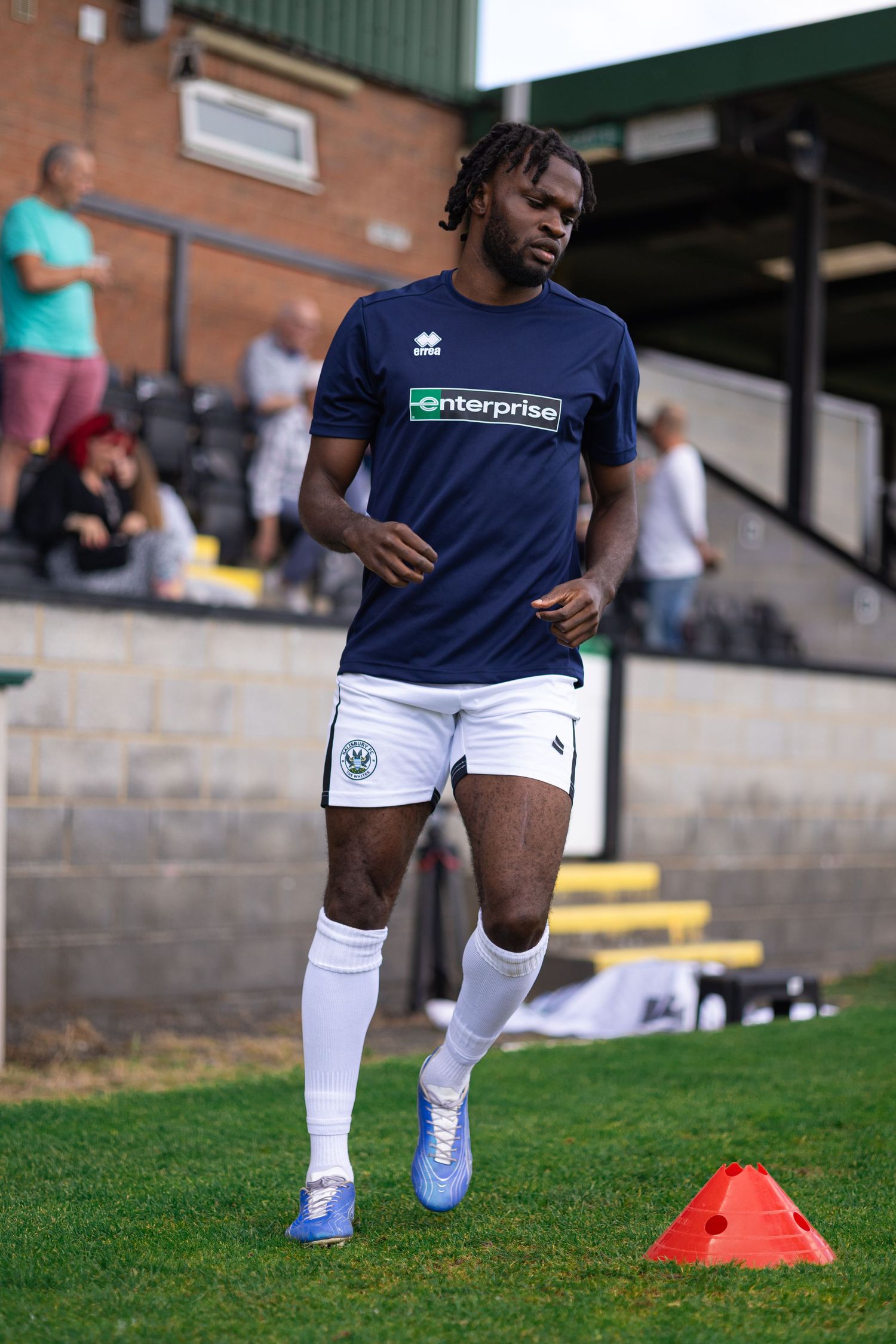 Football player in navy Enterprise-sponsored kit warming up on grass pitch with orange cones during pre-match training.