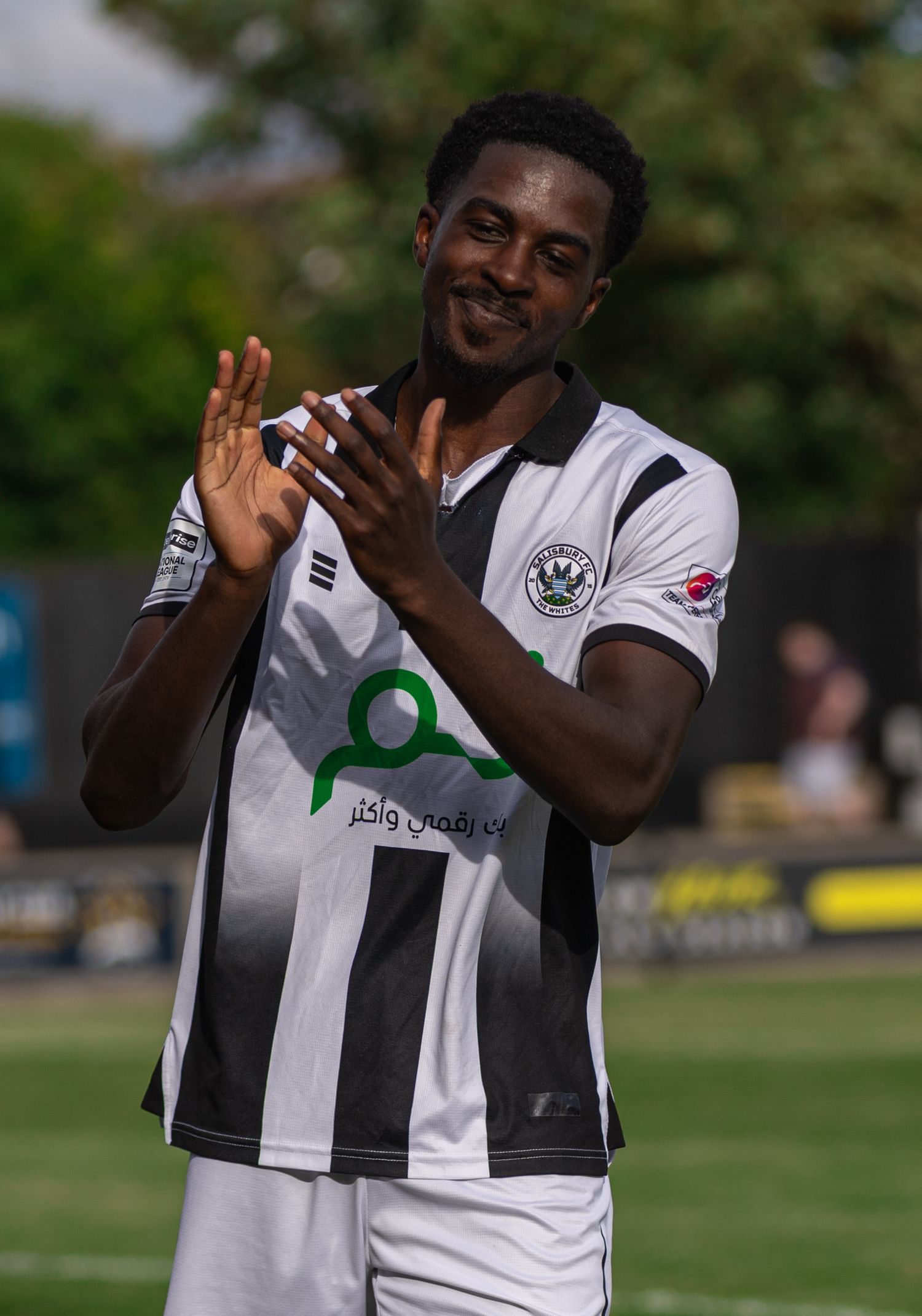 Smiling footballer in black and white striped jersey clapping hands outdoors on a sunny day.