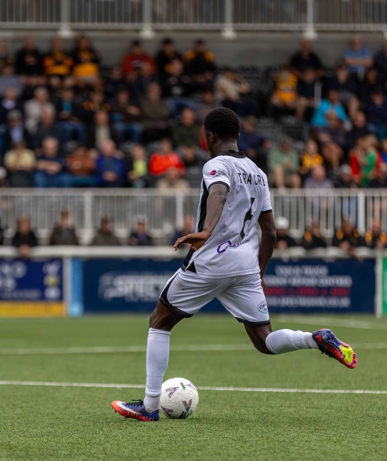 Soccer player number 6 in white uniform dribbles ball on green field with fans in stadium stands behind.