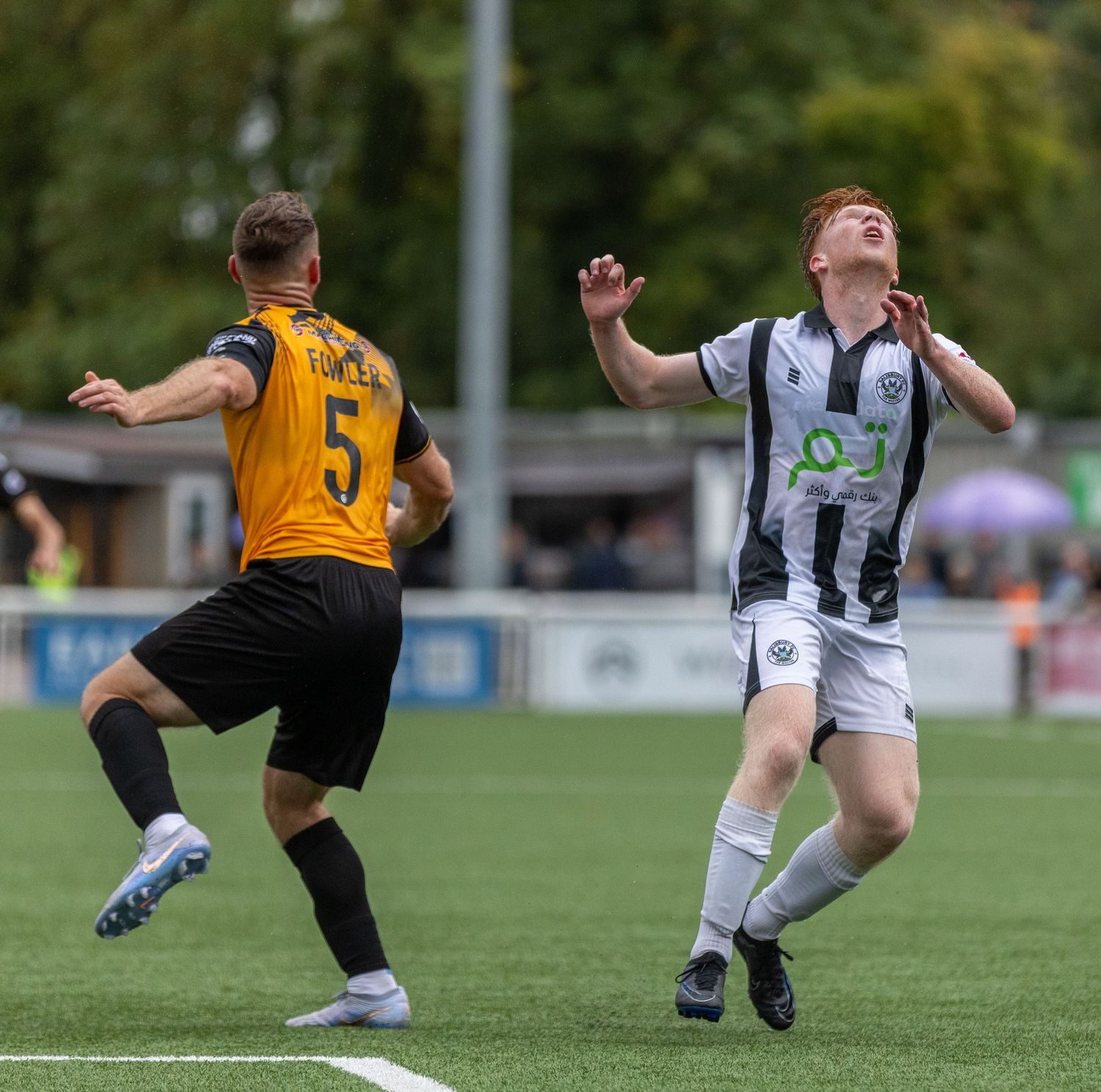 Two soccer players compete for the ball during a match, one wearing gold and black, the other in white and black.